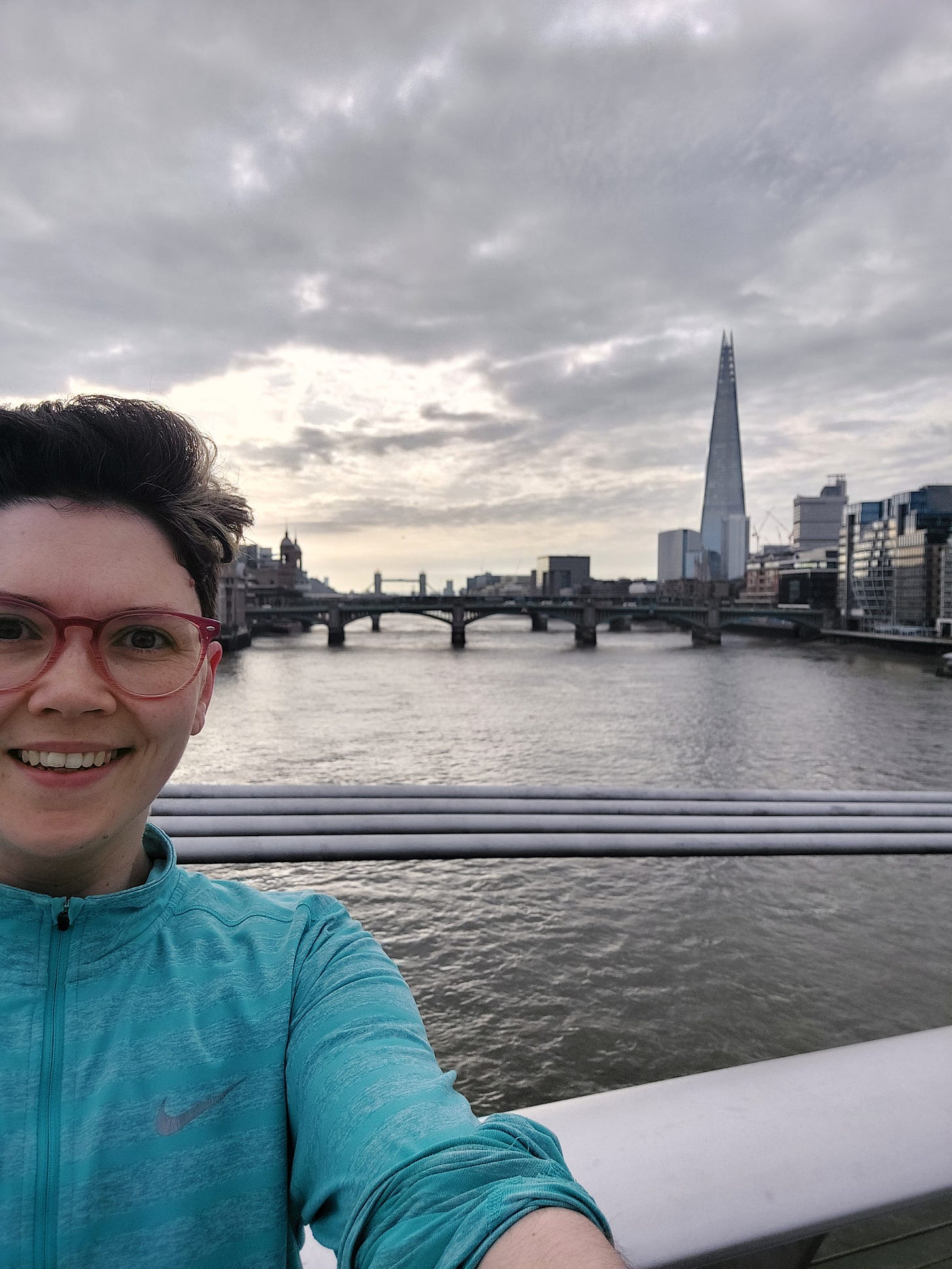 A smiley Janelle (white woman, pink glasses, pixie short brown hair) with the vista of London behind her including the Shard and Tower bridge