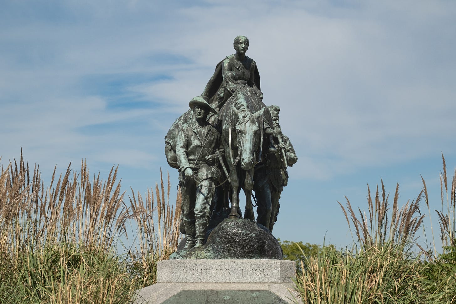 A statue of three white people walking across the prairie: a younger man walking, a woman on a horse, and an older man walking. Set amidst tall grass in a park in Kansas City, Missouri.  titled 