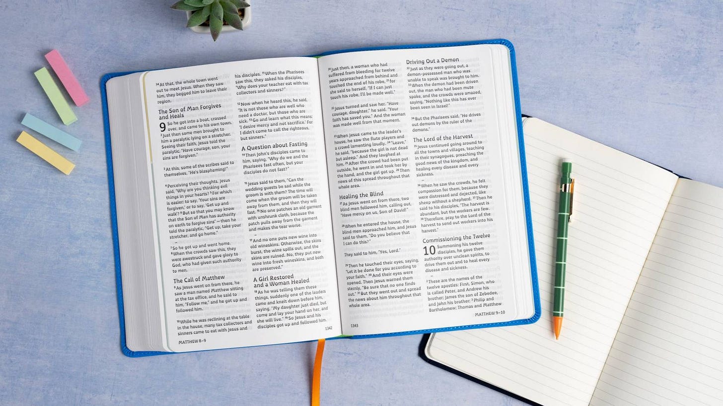 A Bible open next to a blank notebook. there are also sticky notes and pens on the table.