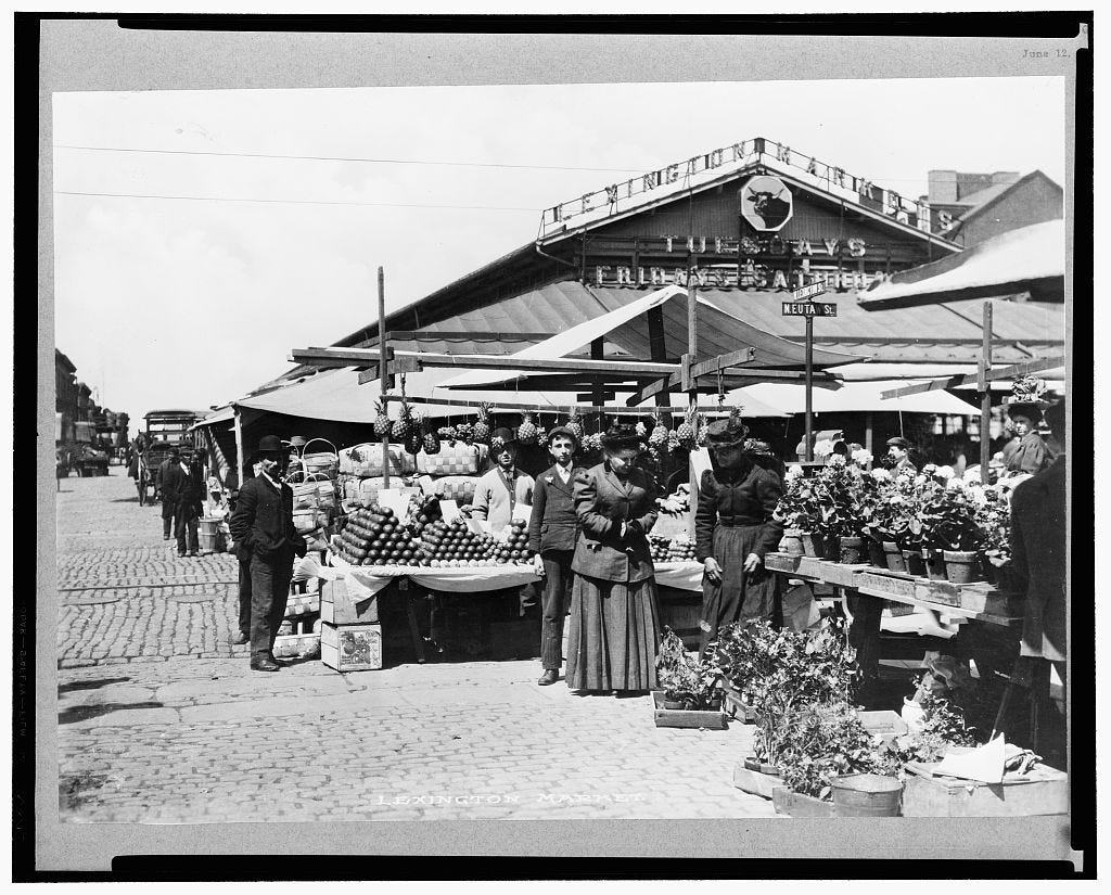 Black-and-white photograph showing Lexington Market, prominently featuring potted plants and stacked produce. Many people interact throughout the market scene.