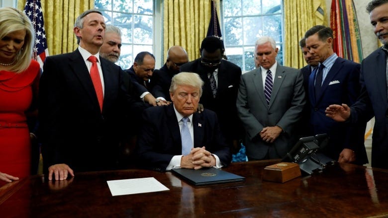 Photo taken in the Oval Office. Trump is seated at his desk, surrounded by many men and one woman, standing, all of them bowing their heads with their eyes closed, as if in prayer. Some of the men have their hands on Trump's shoulders, like disciples of Jesus just touching his robe.