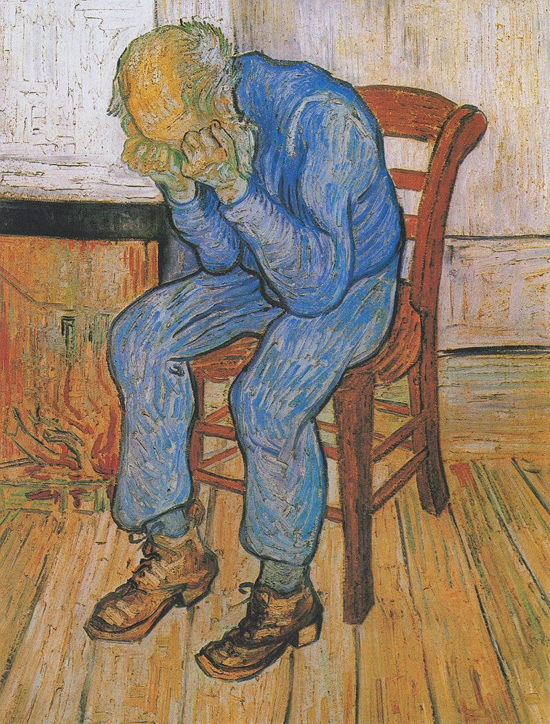 An elderly man with a bald head is sitting on a yellow chair by his fire. There is a low fire in the grate. He is dressed in blue clothes. He is holding his head in his hands.