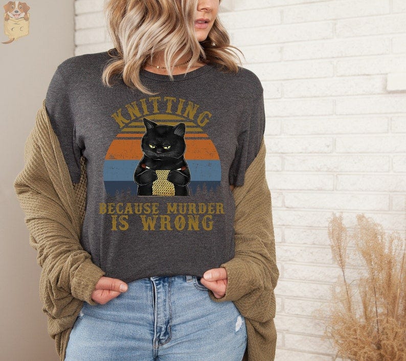May include: A gray t-shirt with a black cat knitting in front of a sunset graphic. The text on the shirt reads 'Knitting because murder is wrong.'