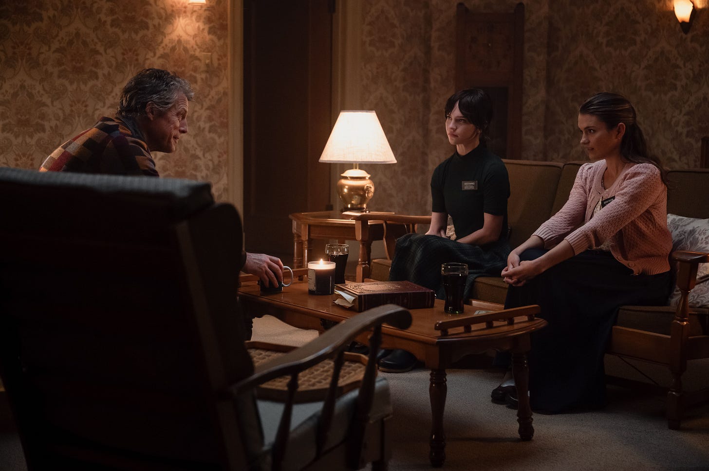 An intense man sits across a coffee able from two female Mormon missionaries in a dim, old-styled living room.