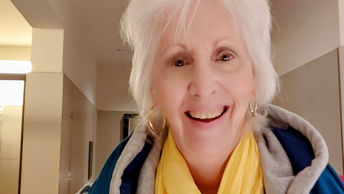 Selfie of Donna Blevins smiling with white hair, yellow scarf. and blue and grey hoodie. Tan background. Gold earrings.