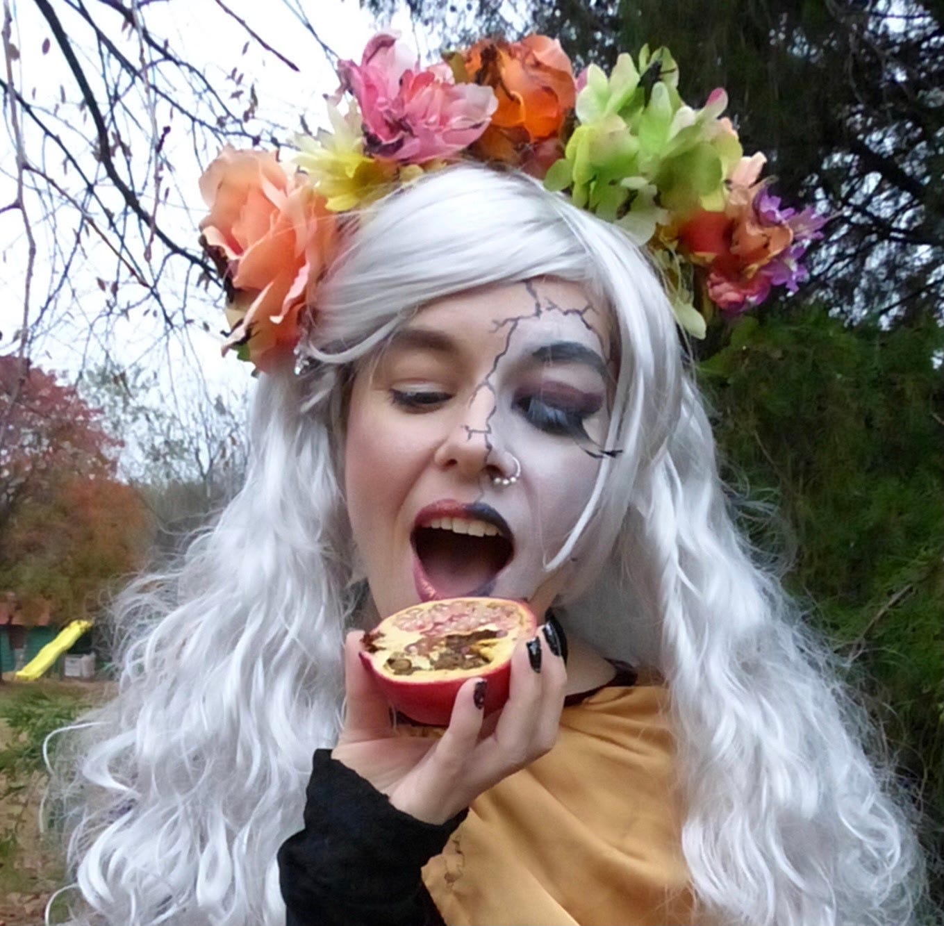 Persephone in a singed flower crown about to take a bite of a pomegranate. Half of it is rotten; half of it is fresh, just like half her face is Blossomtime fresh and the other is the Dread Queen of the Underworld