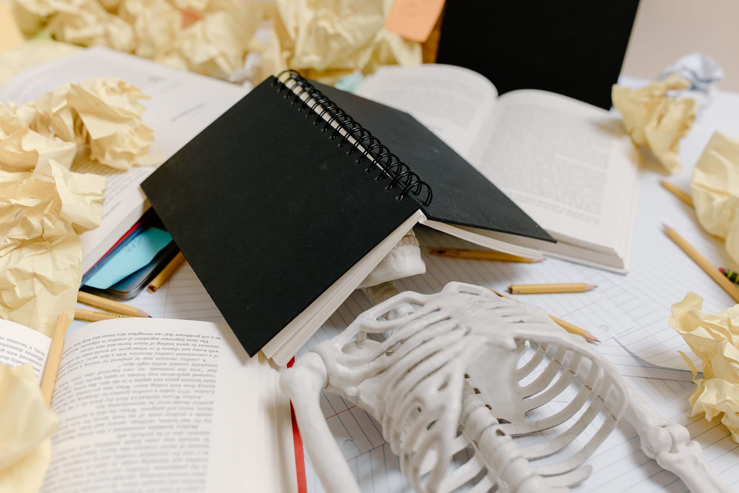 a plastic skeleton is laying on a desk under a notebook surrounded by wadded up yellow papers and writing utensils 