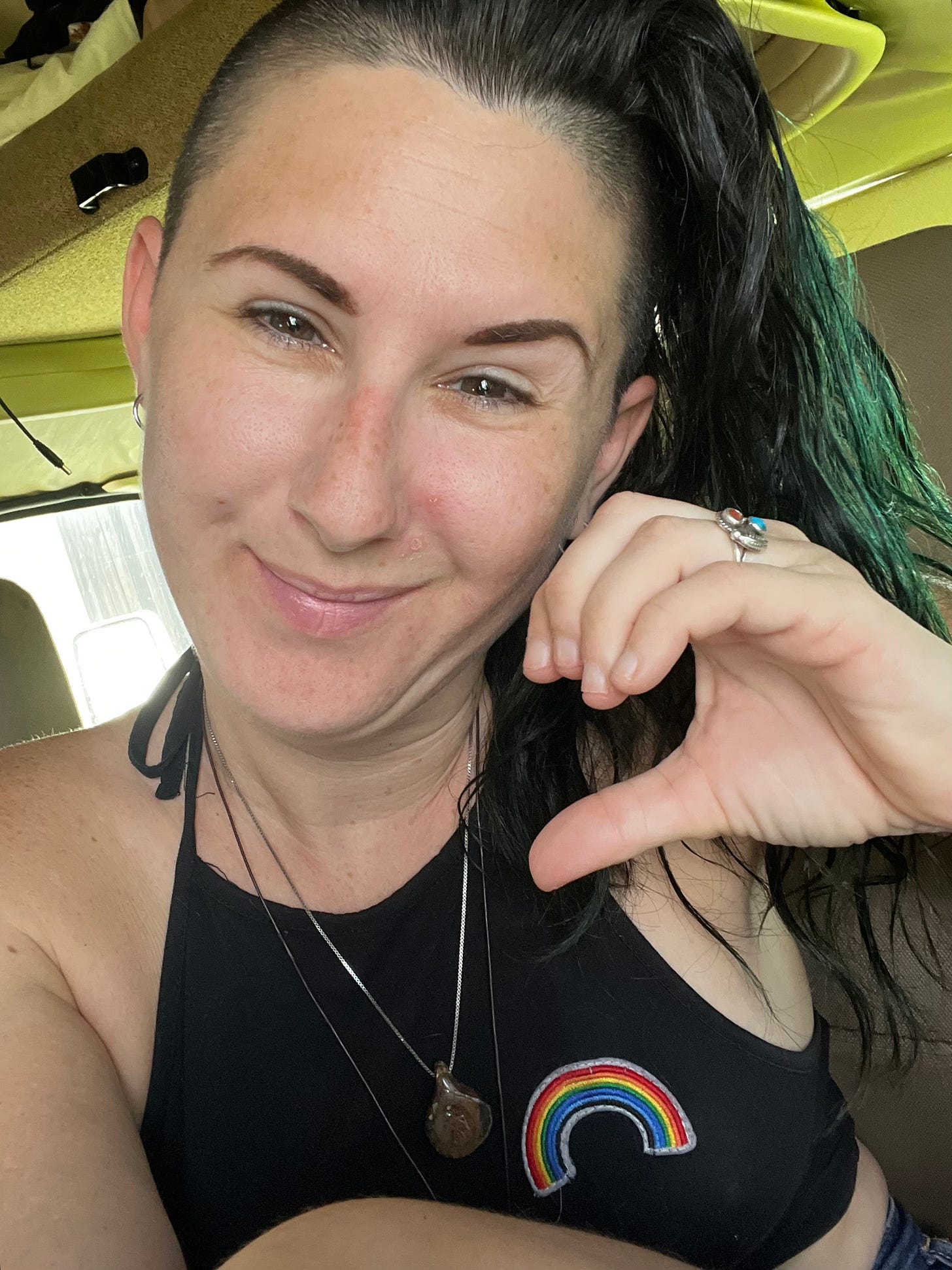 Lyric, more recently, holding up a one half of a heart hand, sitting in their RV smiling. They wear a brown wooden necklace with a thunderbird tack on it, and a black halter top with a rainbow on it, and are smiling at the camerea. 