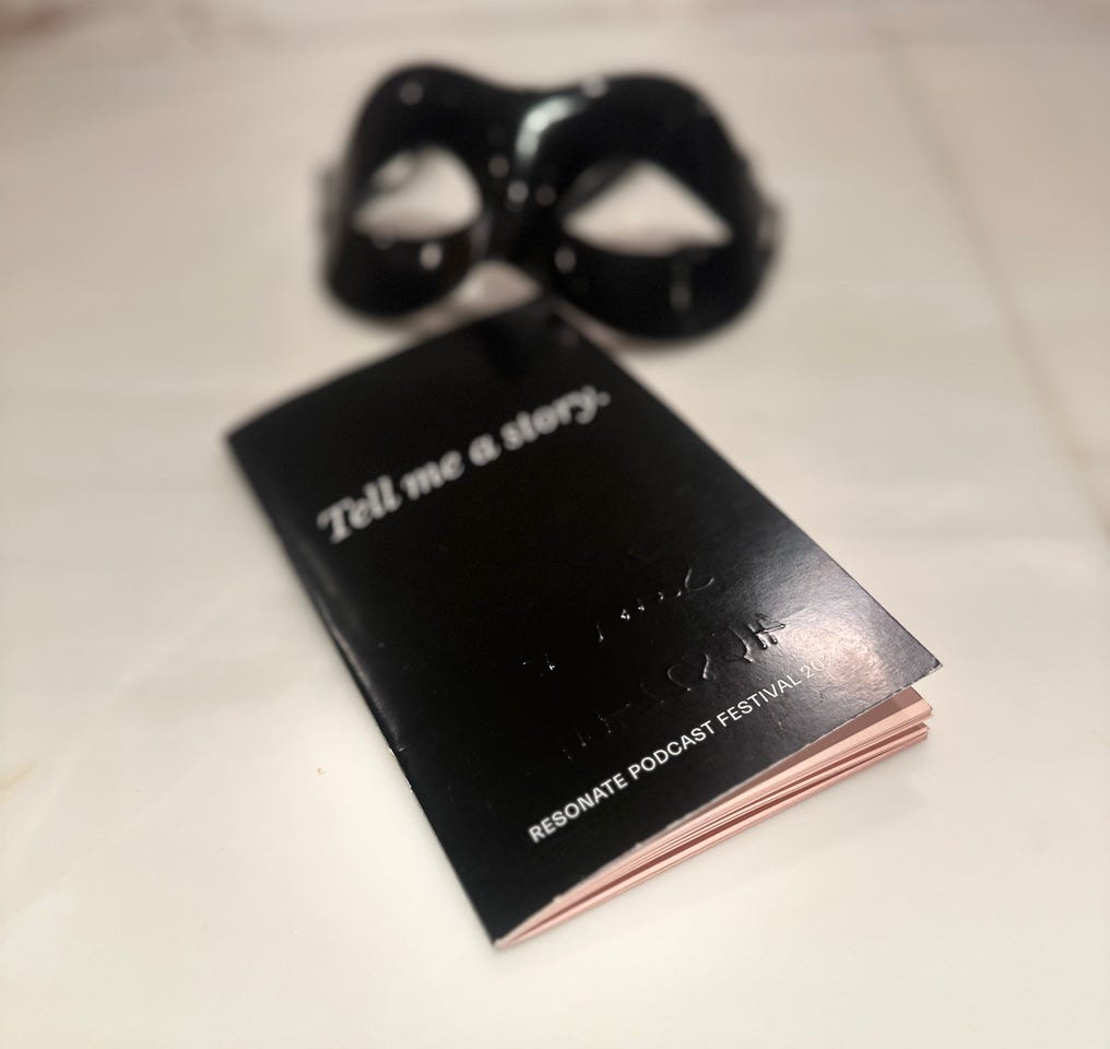 The image shoes a black mask, which is described in the captain as an Eyes Wide Shut mask, next to a black notebook which has the heading Tell Me A Story, and then at the bottom of the small black notebook it says Resonate Podcast Festival 2024