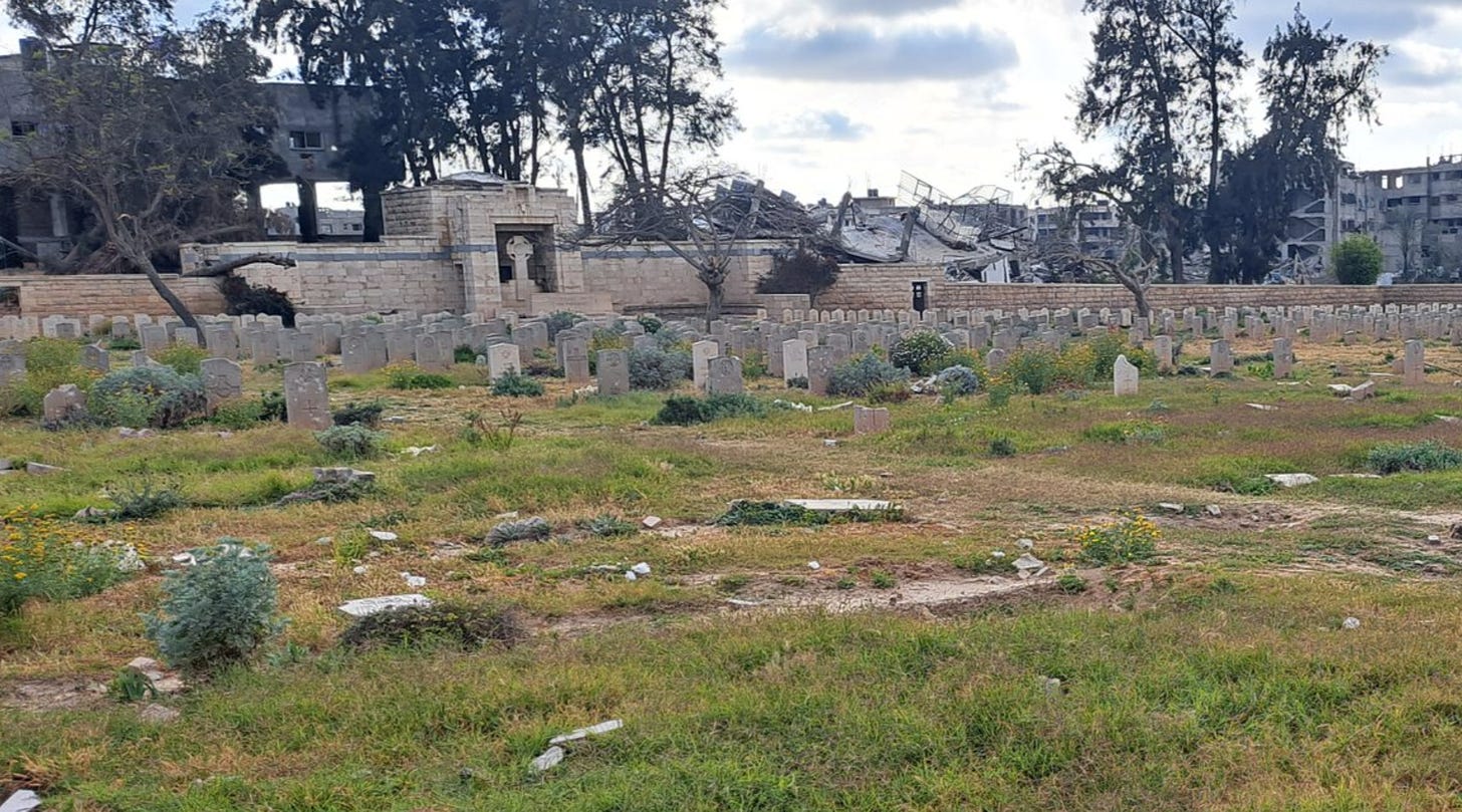 Image of damaged Gaza War Cemetery in early 2024. It shows a view of the central Cross of Sacrifice. In the foreground, several headstones are destroyed, in the middle ground, more still stand. Behind the Cross of Sacrifice structure, destroyed buildings can be seen, one being the CWGC Staff house.