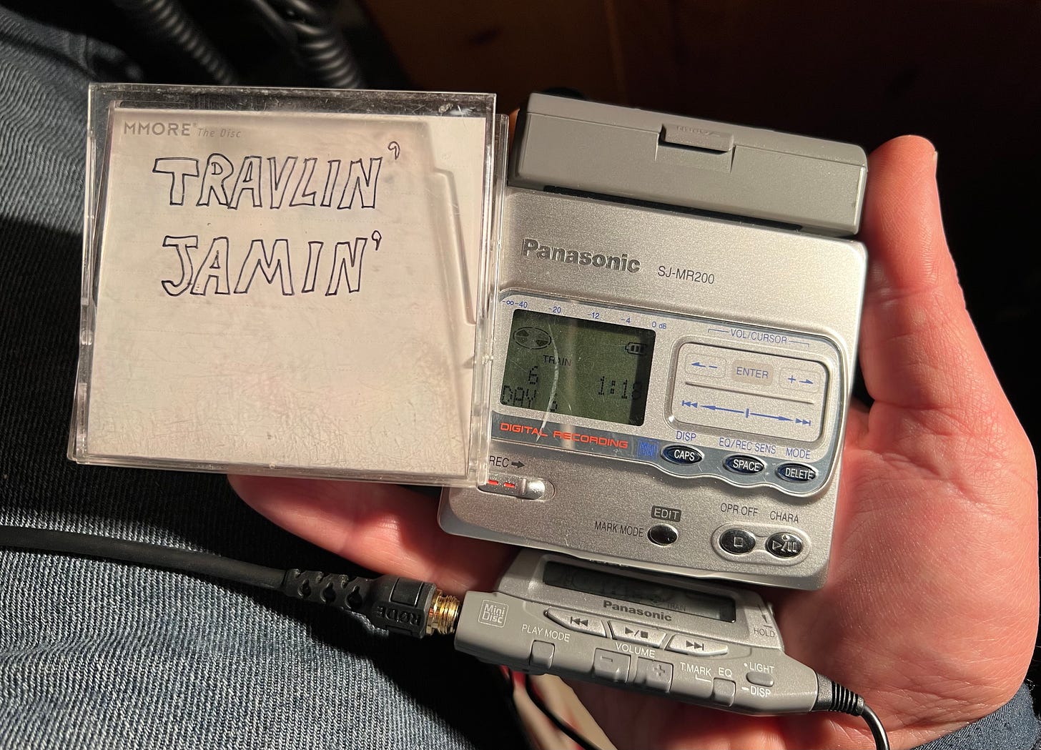 A photograph of my hand holding a mini this case with the words Travlin' Jamin' written in pen to the left of my Panasonic SJ-MR200 mini disc recorder, the MiniDisc contains the music experienced over a few weeks of travel. Mostly played by friends and locals on guitar.