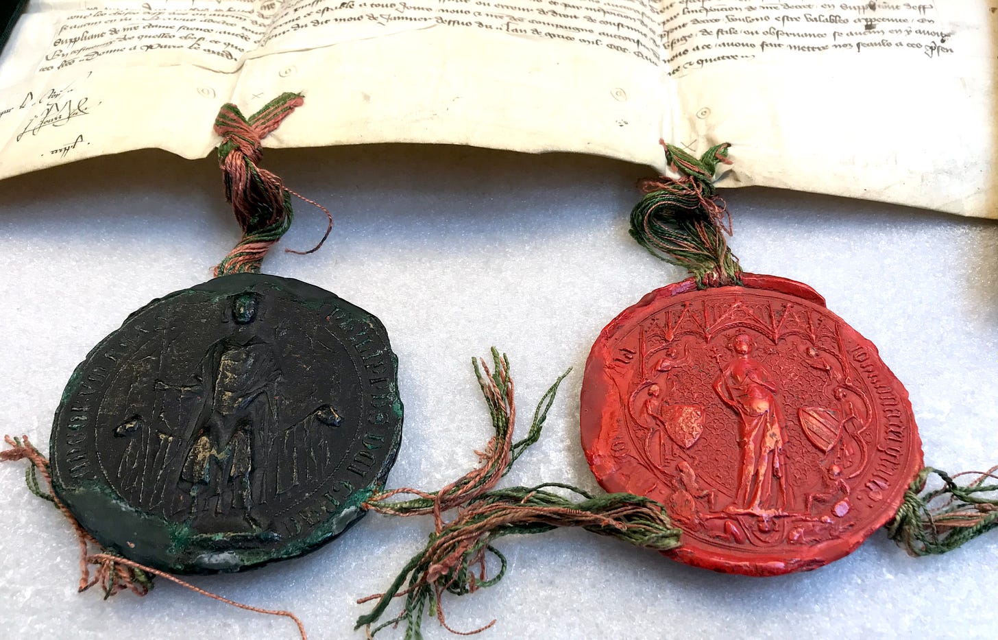 Photo of a fourteenth-century document (partial view), with two large round seals in wax attached to its bottom by silk ties, red and green: first seal to the left is dark green, with a king sitting on a throne holding a scepter; second is a red seal, showing a queen standing holding two small scepters.