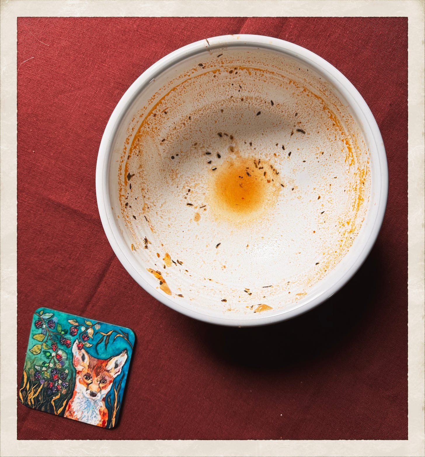 On a red tablecloth is placed a white bowl streaked with herbs and orange stains from food; below that is a colorful coaster featuring a fox.