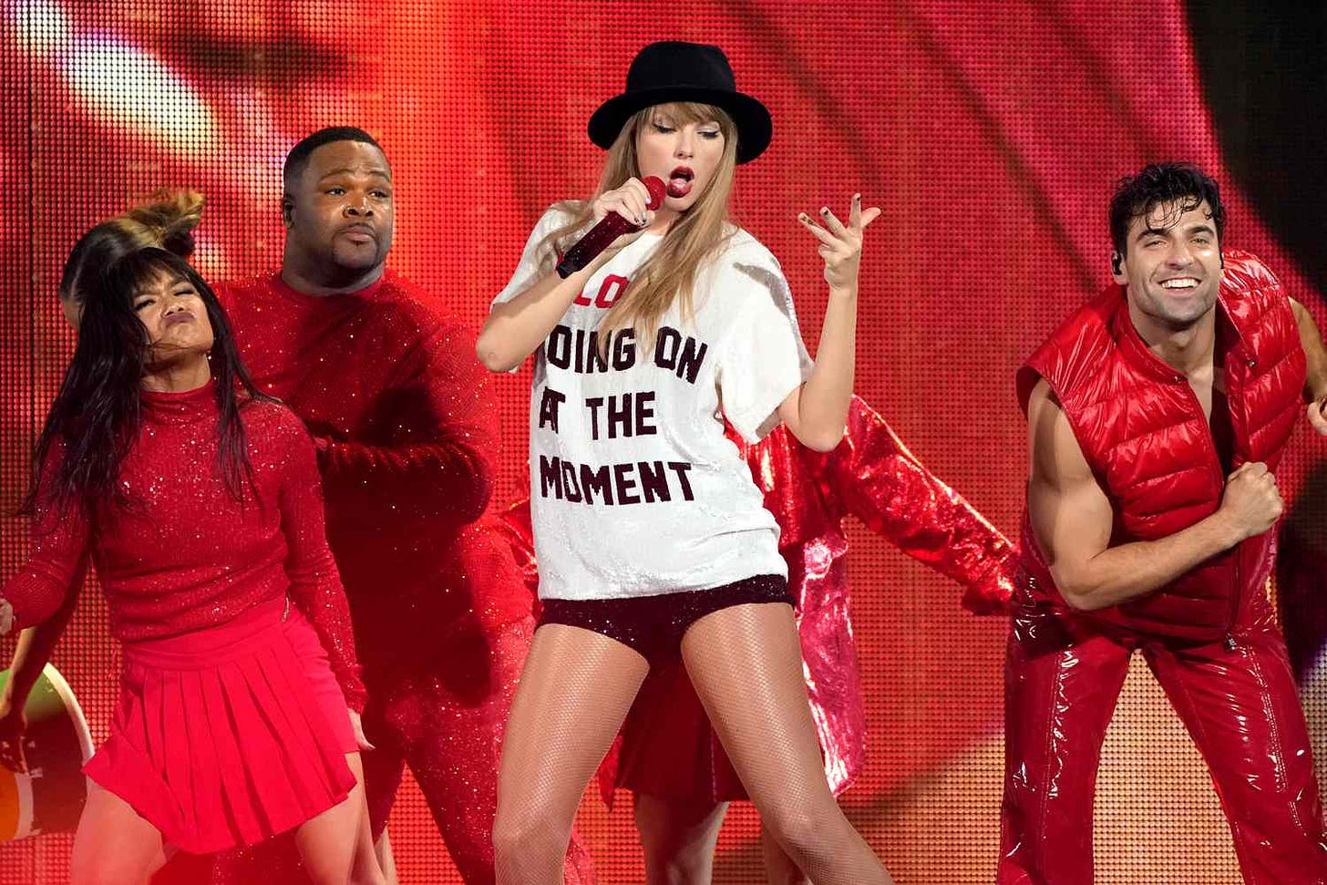 Taylor's Eras Tour red outfit, black hat, white shirt that says "a lot going on at the moment," black mini shorts. Three backup dancers in red outfits, the one on the far left is making a constipated face.
