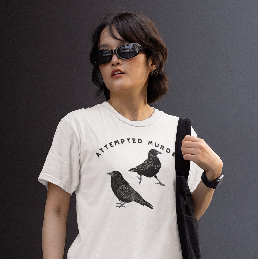 A young woman in sunglasses wearing a white t-shirt with the slogan ATTEMPTED MURDER and illustrations of two black crows.