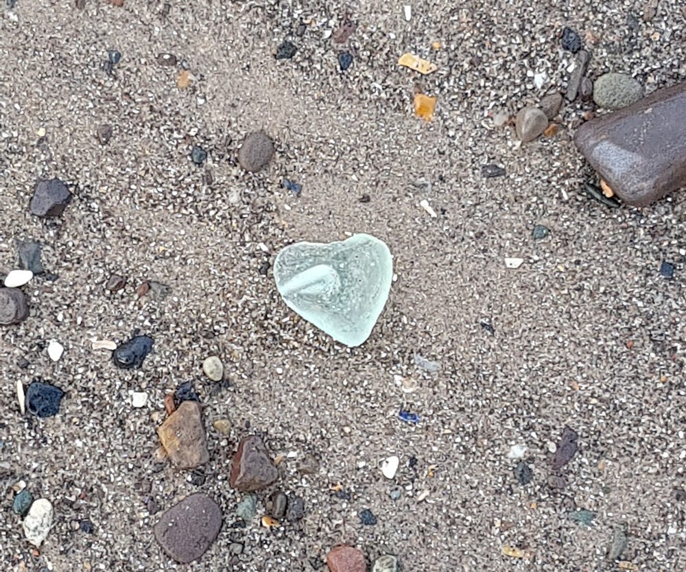 Glowing pale green sea glass heart amid sand and pebbles