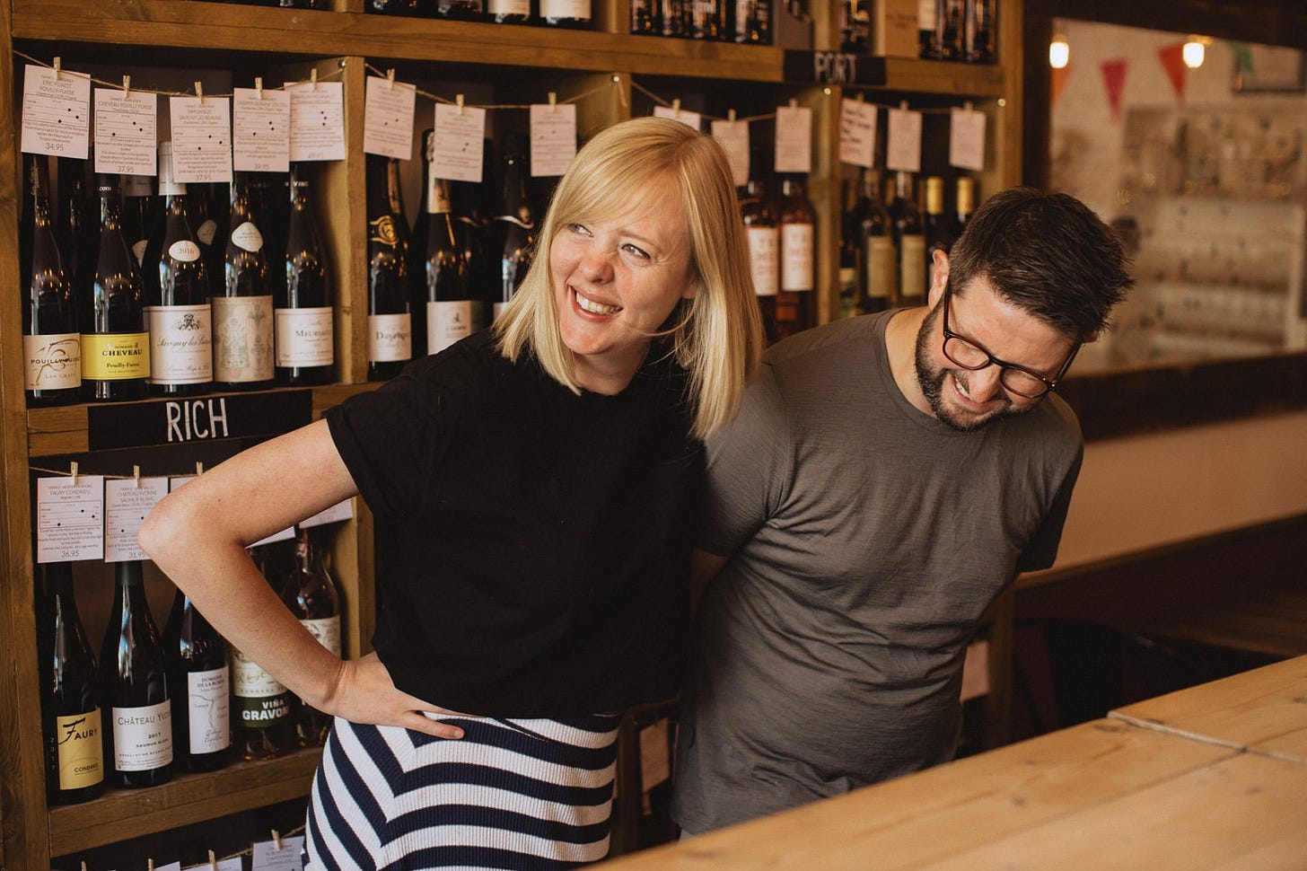 A laughing Charlie and her husband standing in their wine shop, bottles of wine behind them. 