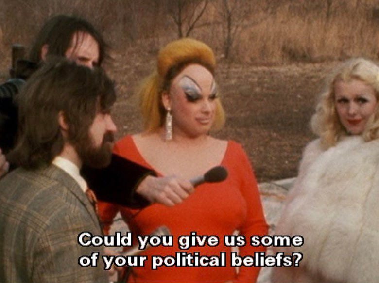 a screen grab from pink flamingos. divine is being interviewed by a reporter. the bottom text reads: "could you give us some of your political beliefs?"