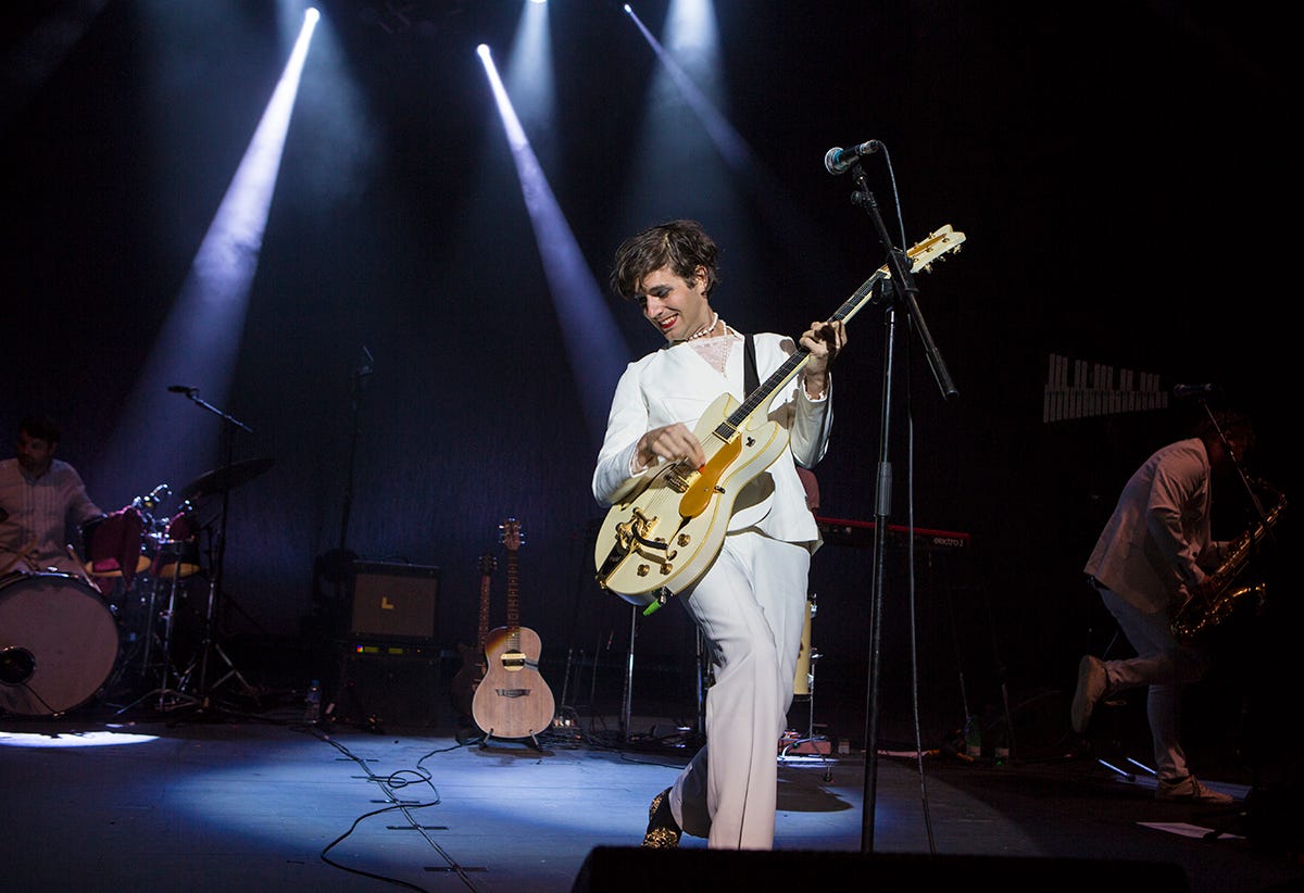Dressed in white with a pearl necklace, singer-songwriter Ezra Furman plays a guitar on stage under streams of white light surrounded by their band.
