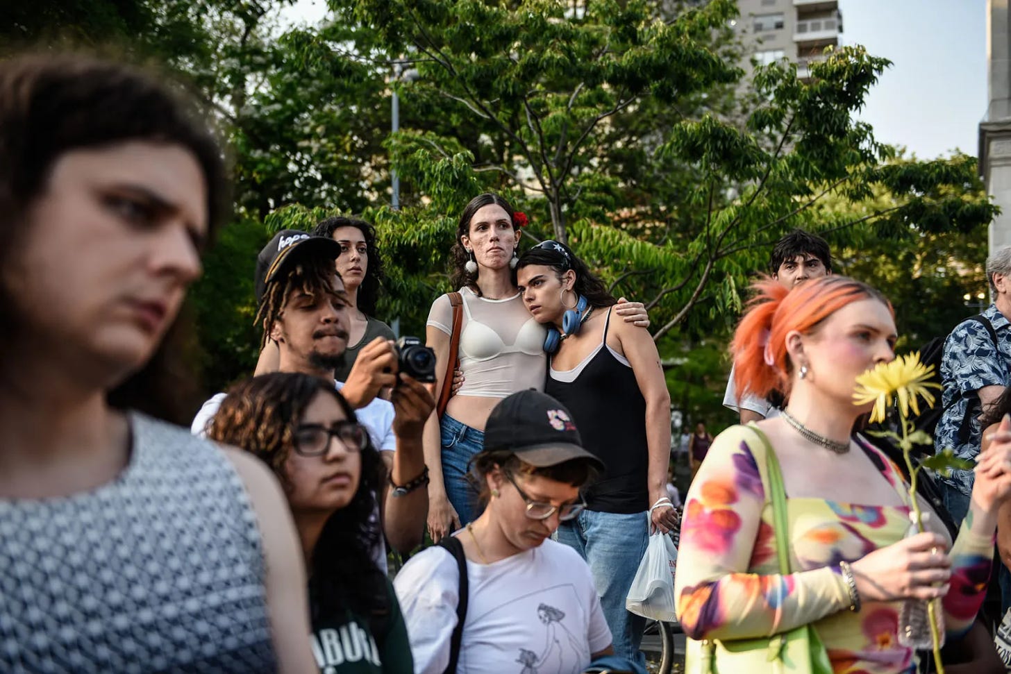 A varied group of people gathered at an event in support of trans people. The group is primarily looking off camera, and some are holding one another. Another holds a yellow flower.