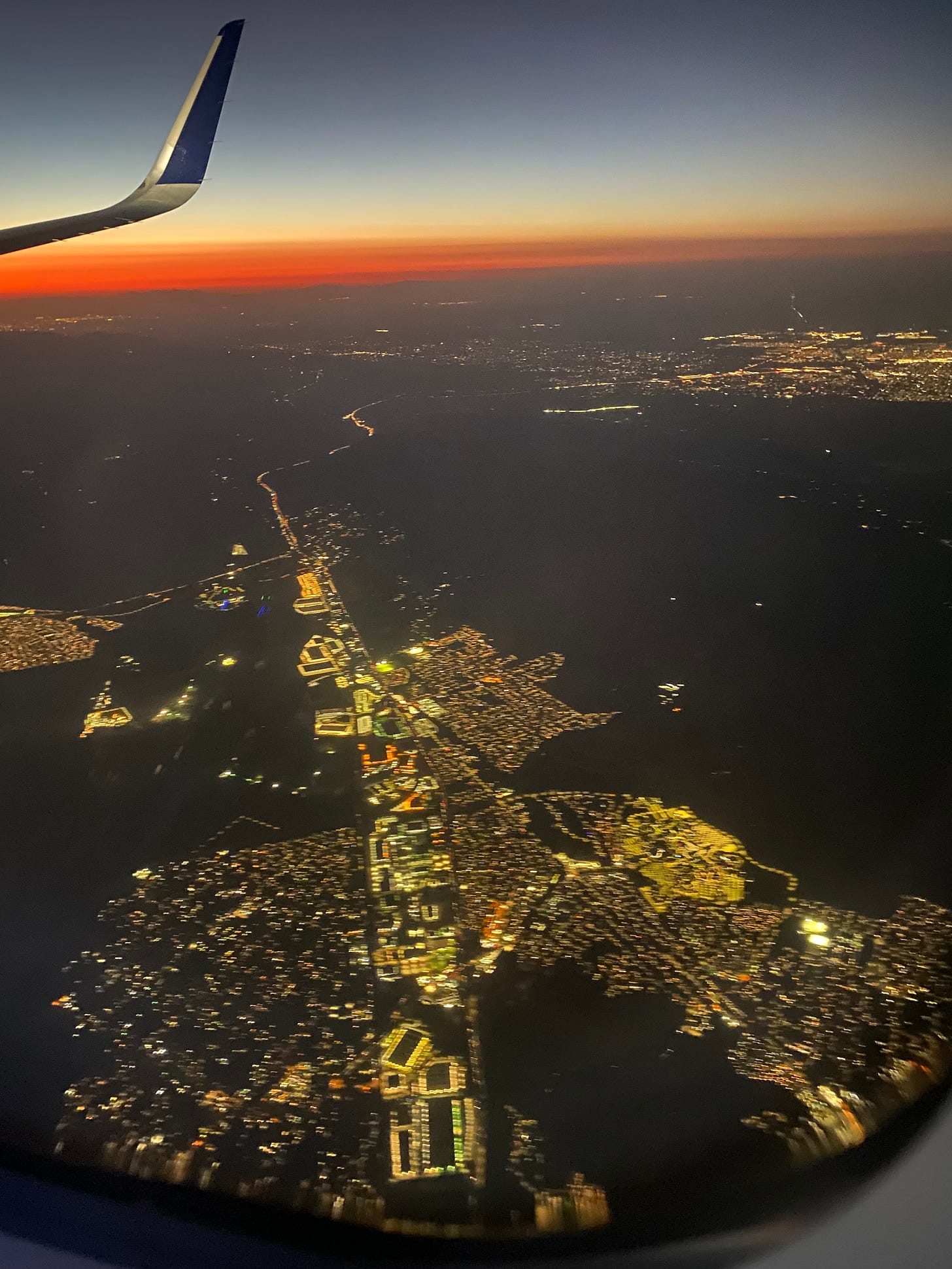 View of LA from an airplane, a red sunset from wildfire smoke and haze, and lights of the city spread out below.