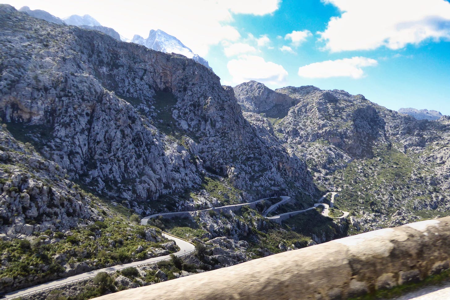 Curving mountain road winding through the rocky Tramuntana range in Mallorca, with steep cliffs and a bright blue sky.