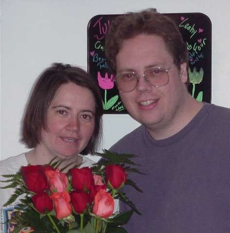 two people in front of a board doodled with tulips and words such as Leahy. Woman holds red and orange roses. both are smiling. man has glasses. woman's hair is messy.