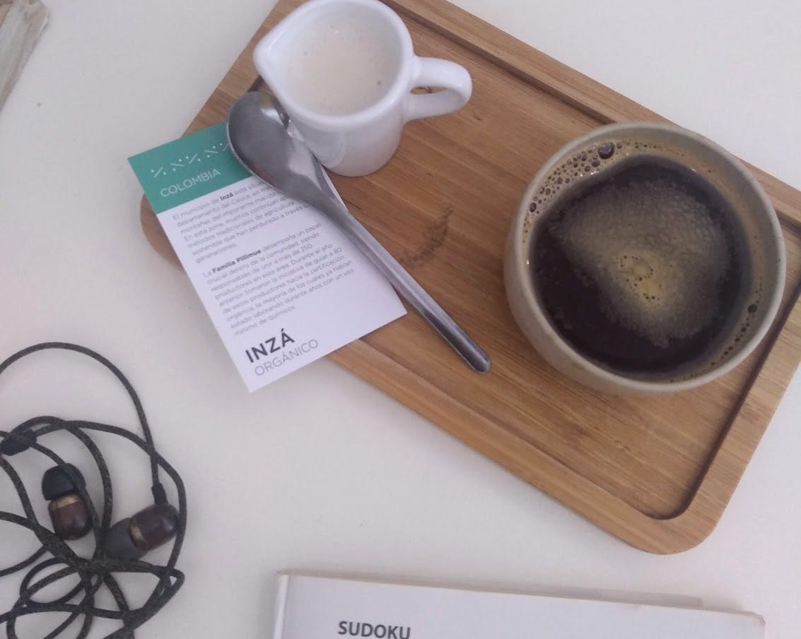 A black coffee on a wooden tray on a white table. There is also a spoon, a small information card on the coffee, and a small pitcher of milk. On the table there are headphones and the edge of a book of sudoku.