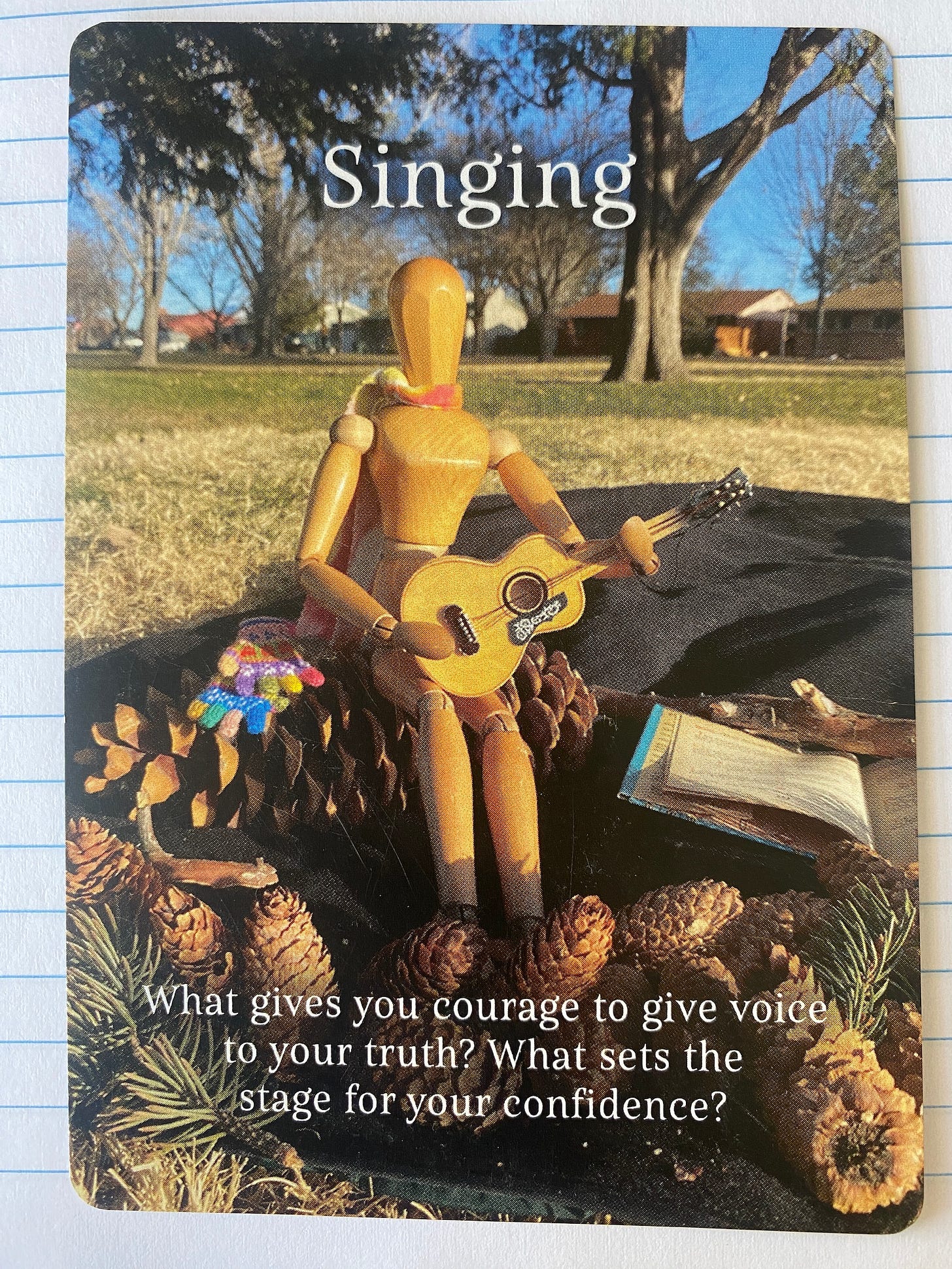 Supposing Card with the Emotikin art manikin (inner artist) sitting on a picnic blanket holding a guitar and pinecones gathered on the blanket. The card is titled Singing and the caption says What gives you courage to give voice to your truth? What sets the stage for your confidence?"