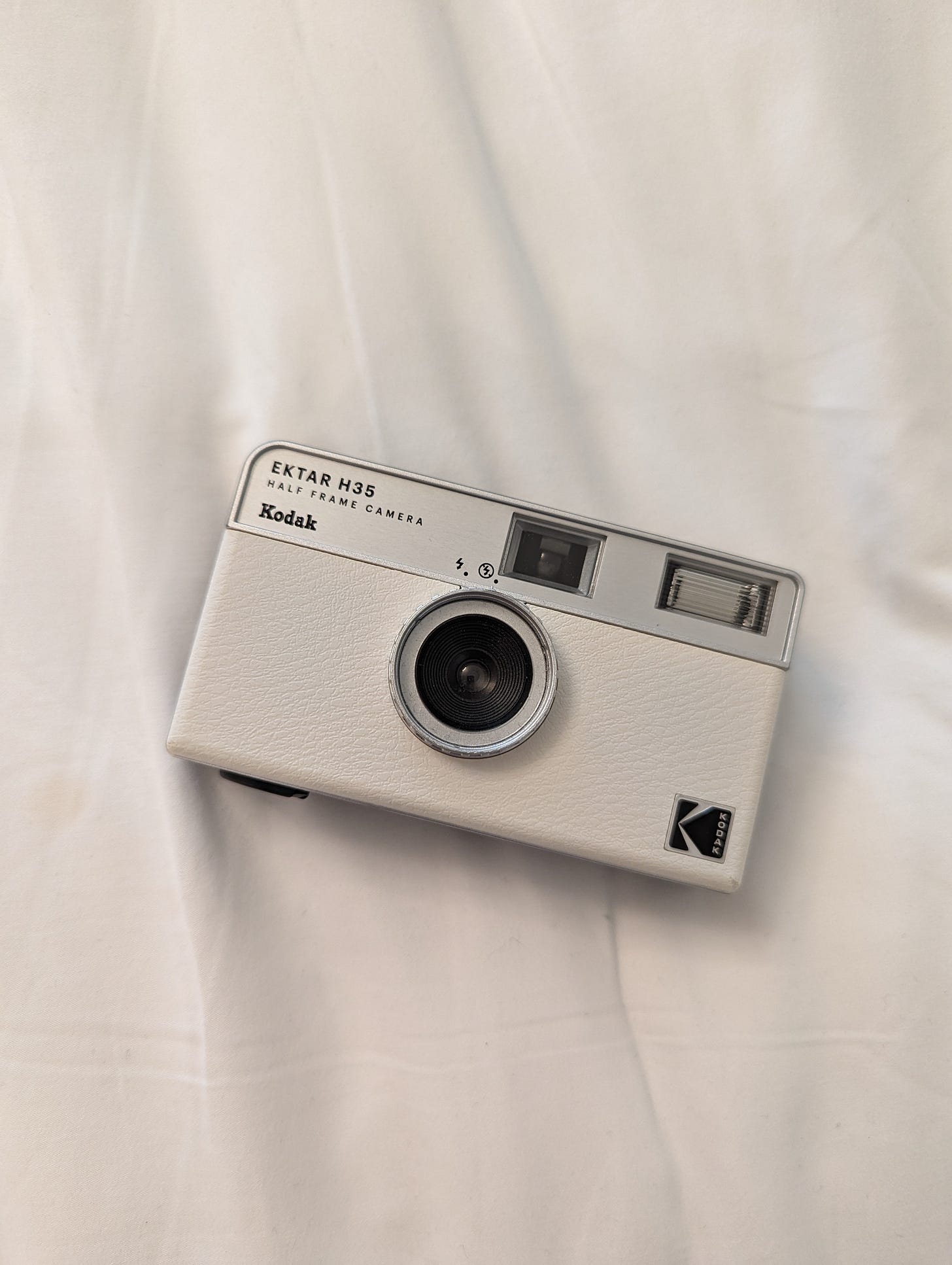 Photo of a Kodak Ektar H35 Half Frame Camera that's white and sits on the white linen of a bedsheet