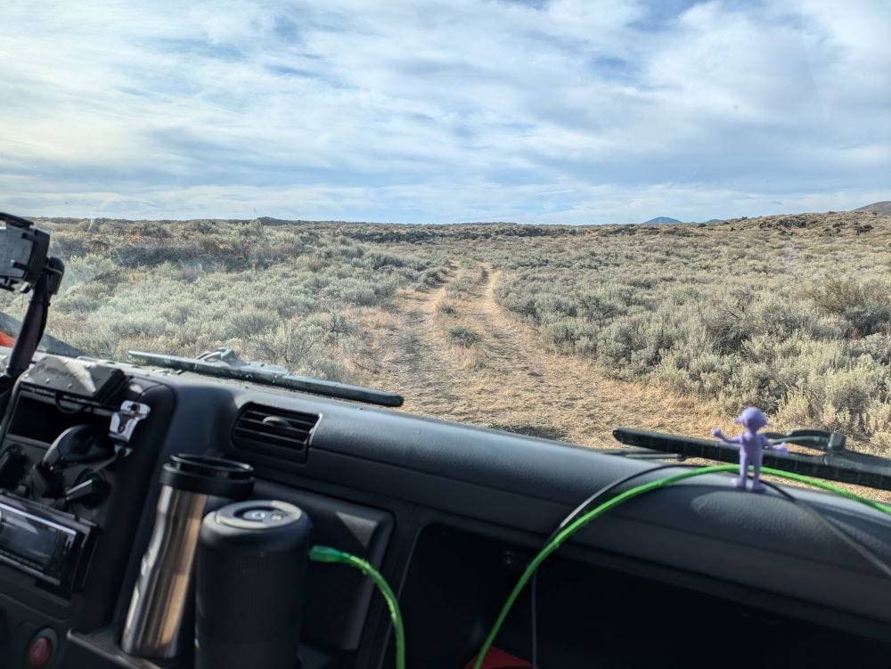 Looking out the windshield of a big truck onto a small dirt road, just two tire tracks across a desert of sagebrush covered hills