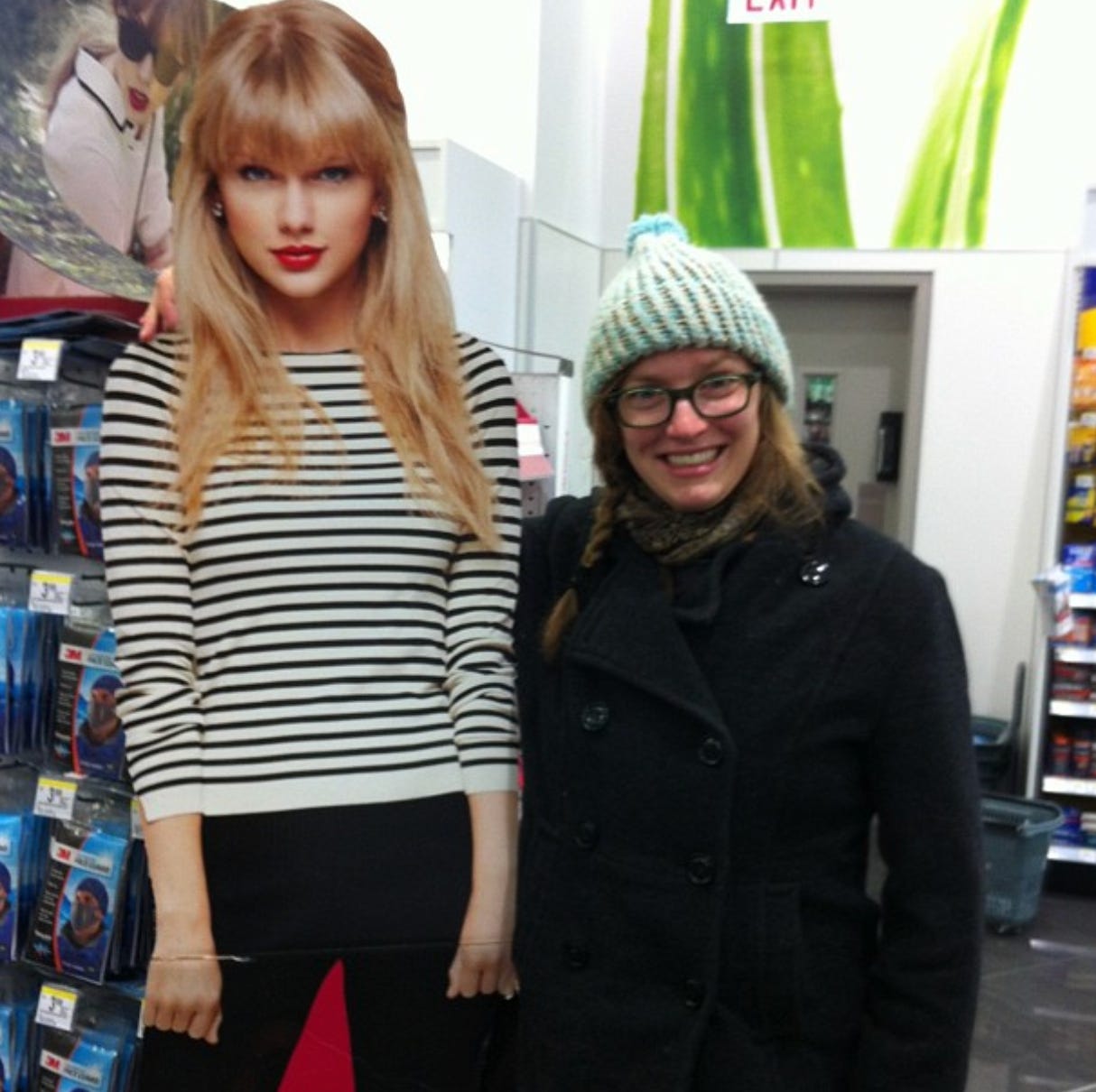 A cardboard cutout of Taylor Swift circa 2012, standing about 6 feet tall with straight hair, lipstick, and bangs. torri stands next to her in a wooly knit hat, chunky jacket, and frizzy hair in braid. 
