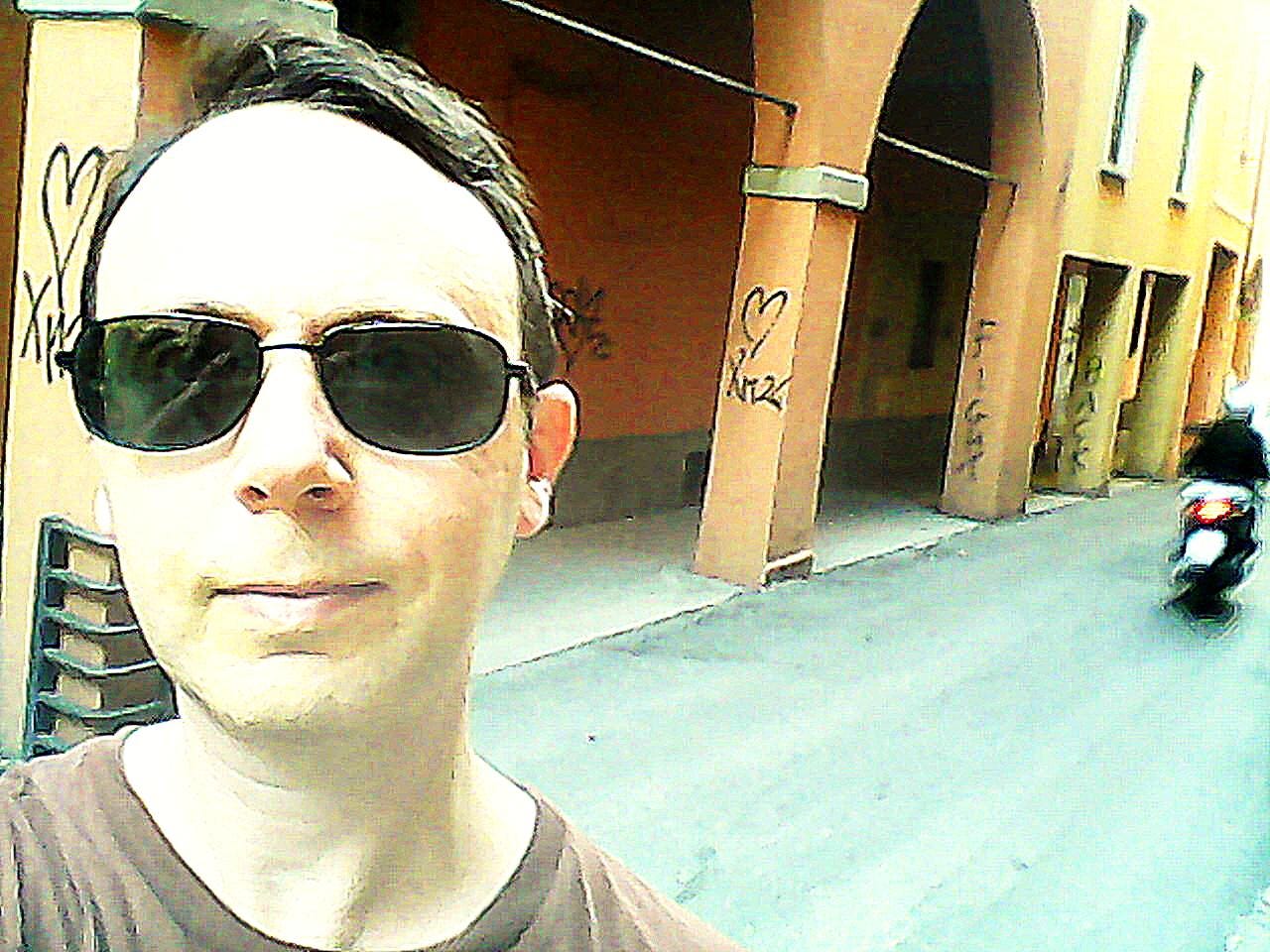 Portrait of a white male with sunglasses with a street in Bologna in the background. Orange and yellow columns have graffiti reading "Fight back."