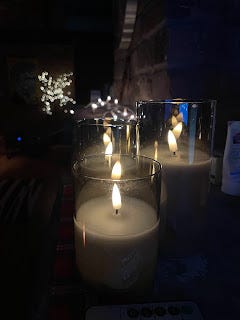 It's a close up of a mantlepiece made of bricks, in a darkened room. There are three lit candles on the mantlepiece, and a string of fairy lights across the length of it. All the lights are a soft white glow in the darkness.