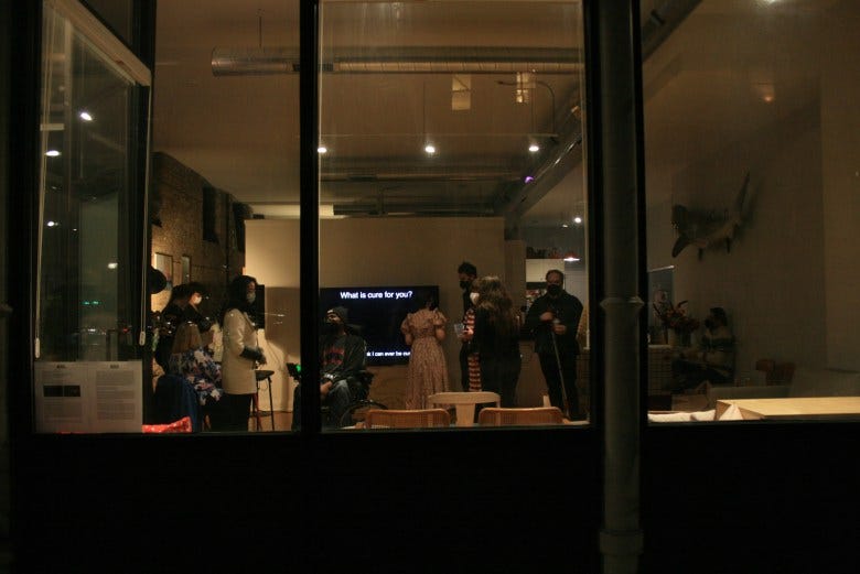 A photo from outside an exhibition space behind glass windows where people mill about at an opening.