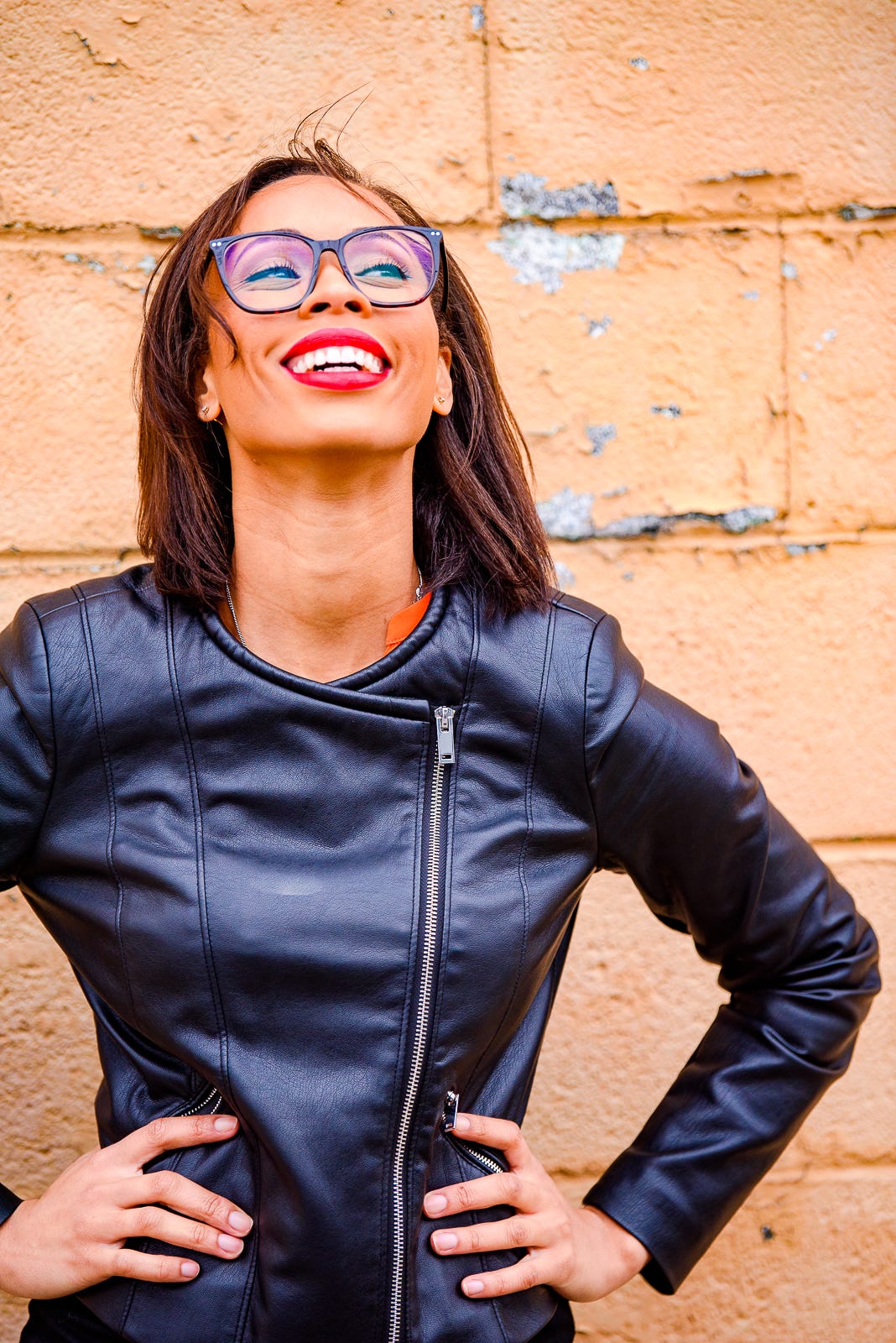 photograph of India Tizol (formerly Jackson) laughing outside against a concrete wall. India is an african american woman with medium length brown hair, brown eyes and big rectangular glasses. She wears a vegan leather jacket (black) and red 999 christian dior liptstick. it is fall/winter in virginia beach, va, USA