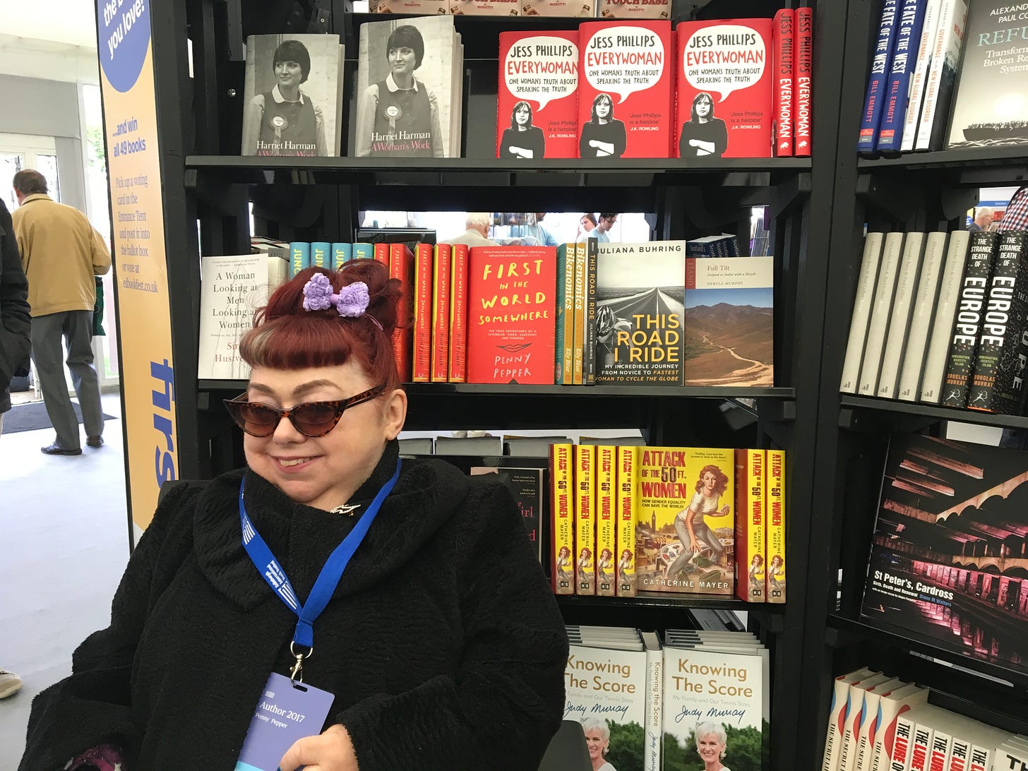 Penny sits smiling in her wheelchair in front of books in a bookshop. There are many rows of shelves with books, and different colours on their front covers. To the right of Penny and behind her head are copies of her memoir, First in the World Somewhere, a red book with yellow lettering. Penny wears black cat’s eyes sunglasses, a black jacket with a purple bow in her red hair. As an author, she wears a blue lanyard (an identity tag), around the neck for the Edinburgh International Book Fair 2017.