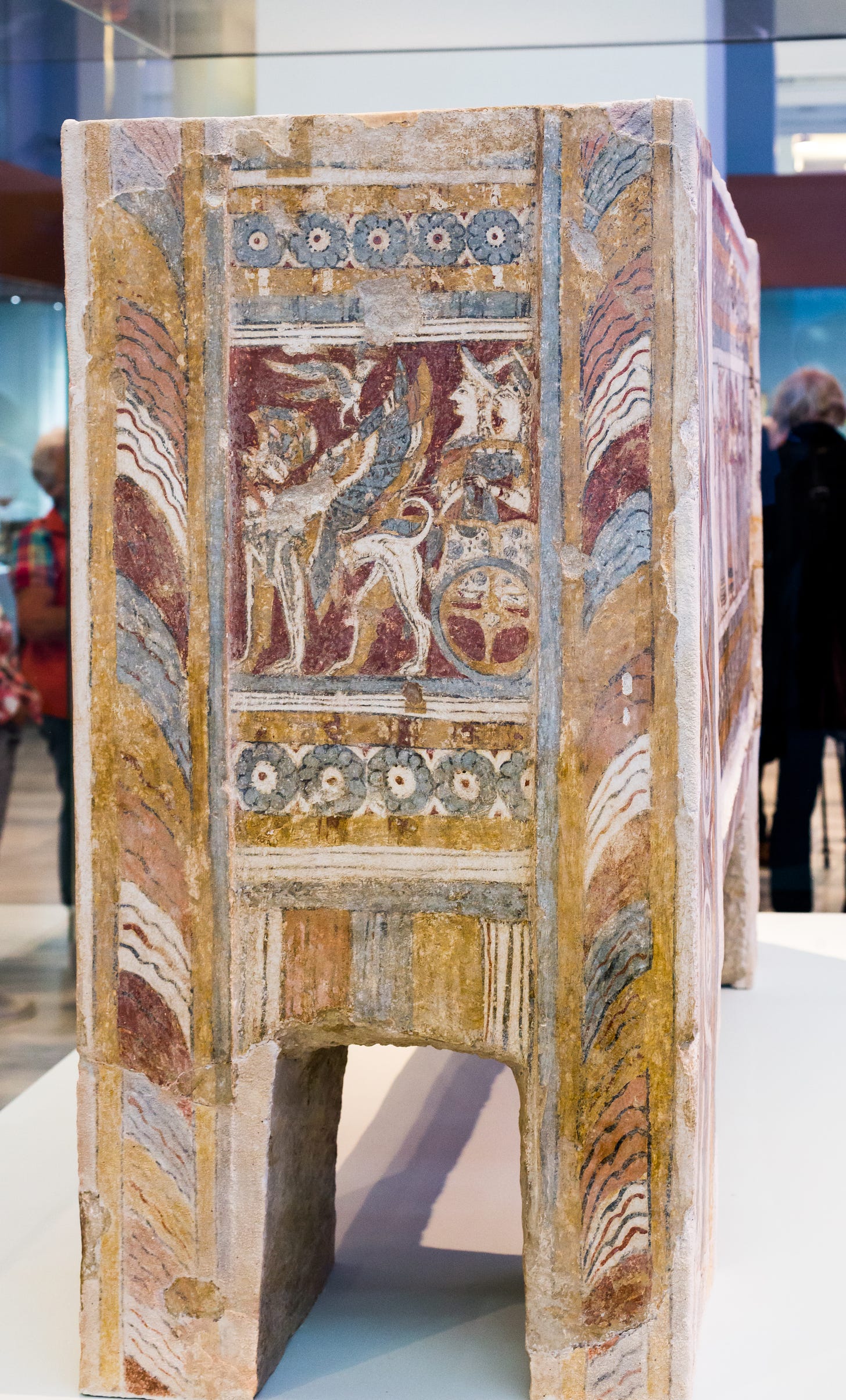 One of the narrow ends of an ancient Minoan sarcophagus, painted with an image of two goddesses riding in a chariot that's being pulled by a griffin. A bird flies above the griffin.