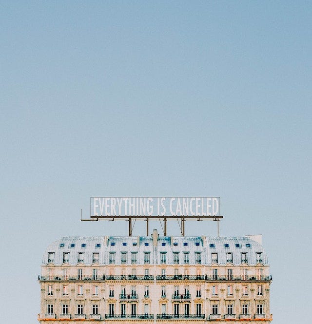 Against a faded blue sky, a large mansion/hotel, with a sign atop saying “Everything is cancelled”.