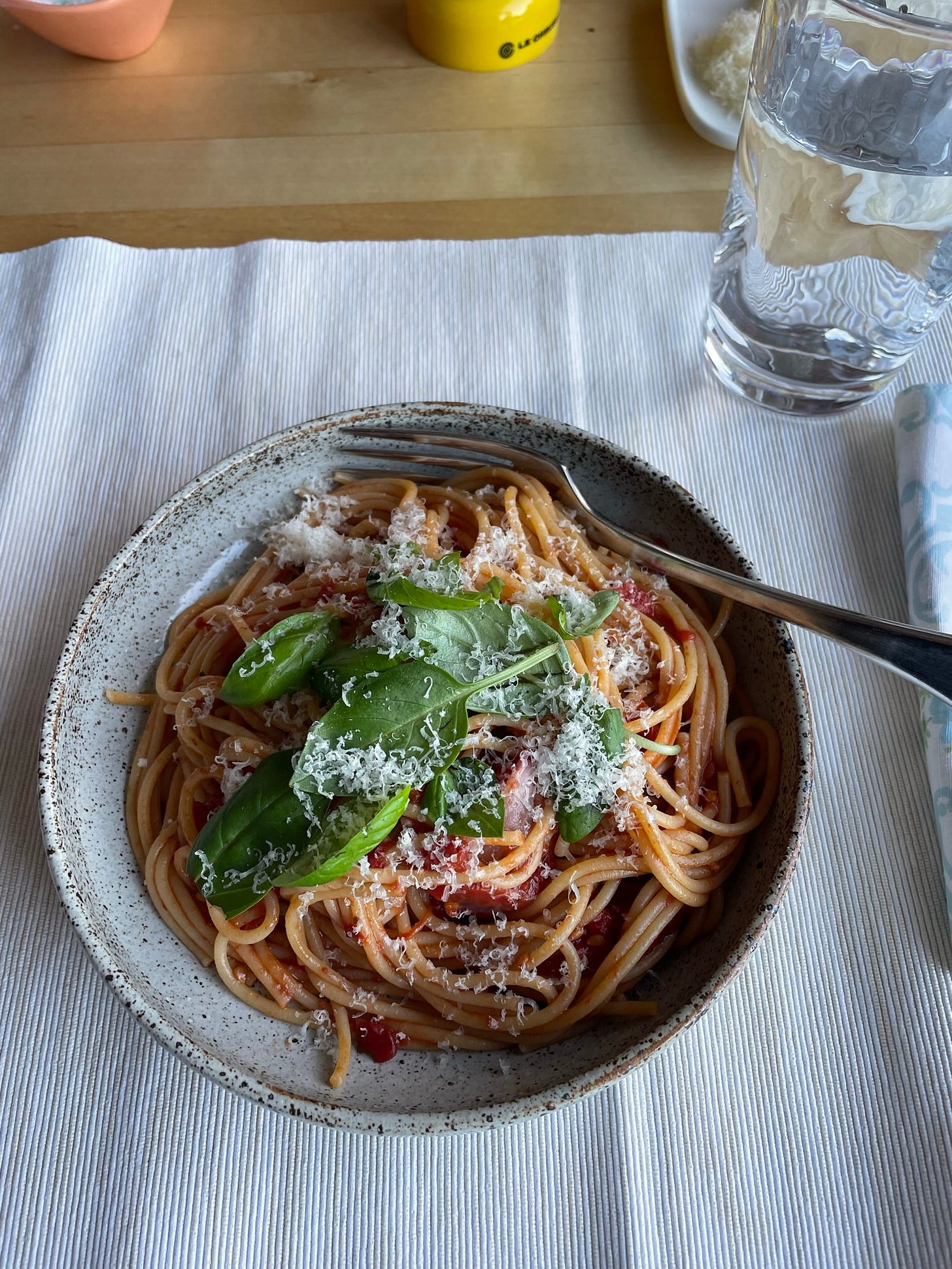 Grey speckled bowl of tomato pasta topped with grated Parmesan cheese and fresh basil leaves. On the dining table is a glass of water, cream placemat, napkin, salt and pepper.