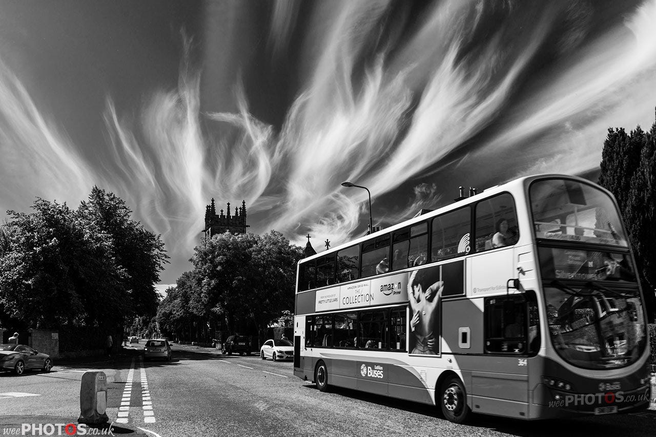 a bus drives past a church steeple with a dramatic wispy octopus-like cloud waving in the sky above