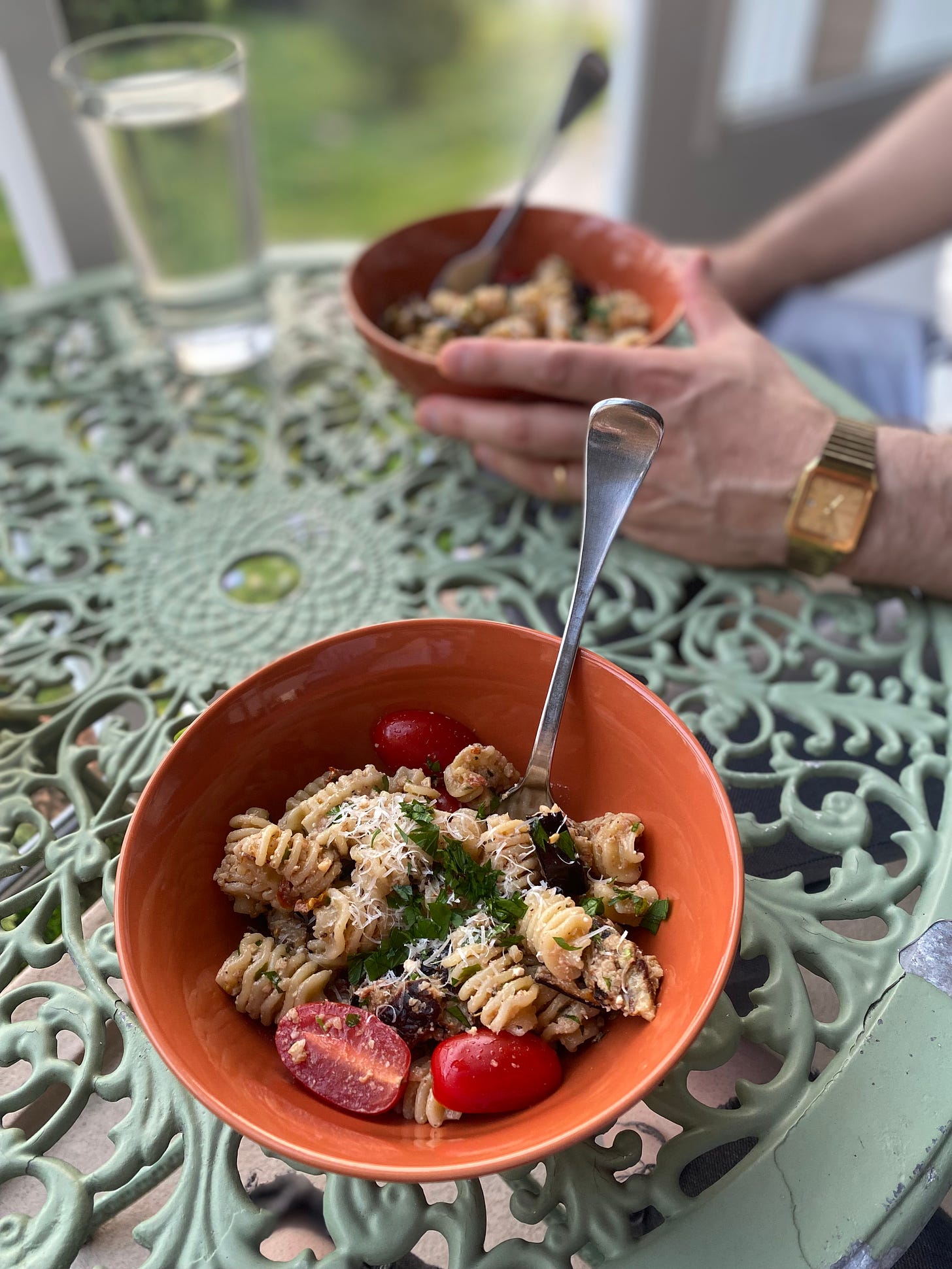 On an outdoor café table, two orange bowls of the pasta salad described above: radiatore with grilled eggplant pieces and grape tomato halves. Everything is dusted with parmesan and parsley. Jeff's hands are visible near his bowl in the background.