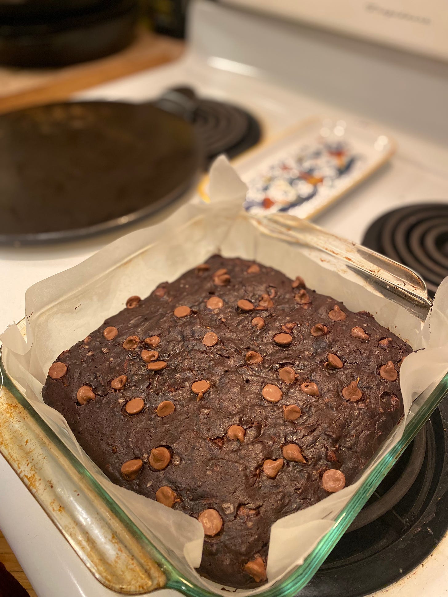 On top of the stove, a square glass baking dish lined with parchment, holding dark brownies dotted with milk chocolate chips.
