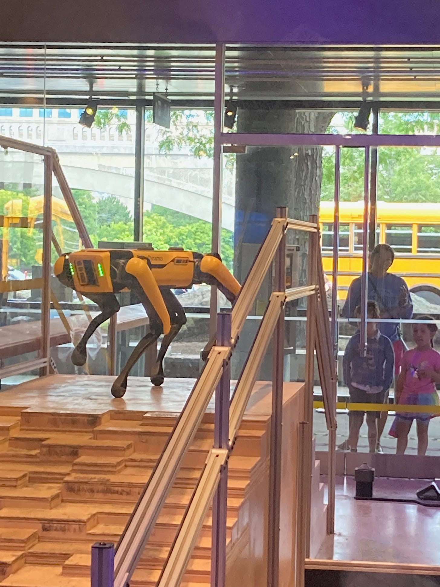 A yellow and black robot dog at the top of stairs with kids looking through a window on the other side
