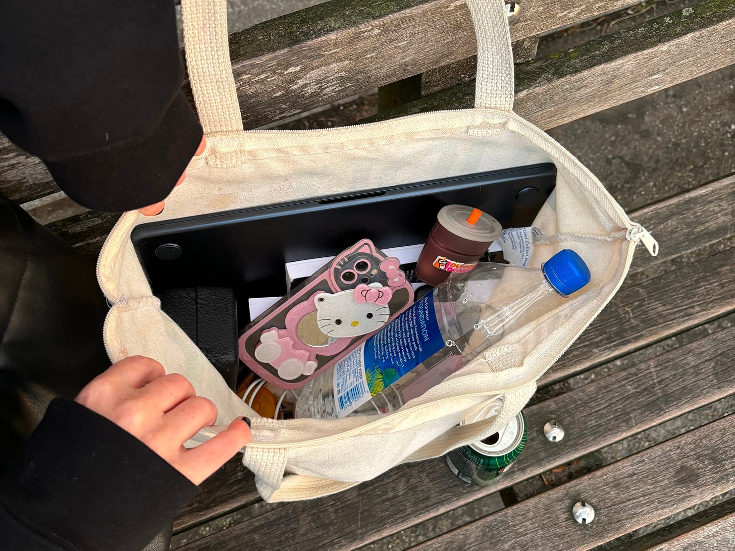 A canvas tote bag rests on a wooden park bench, open to show the items inside