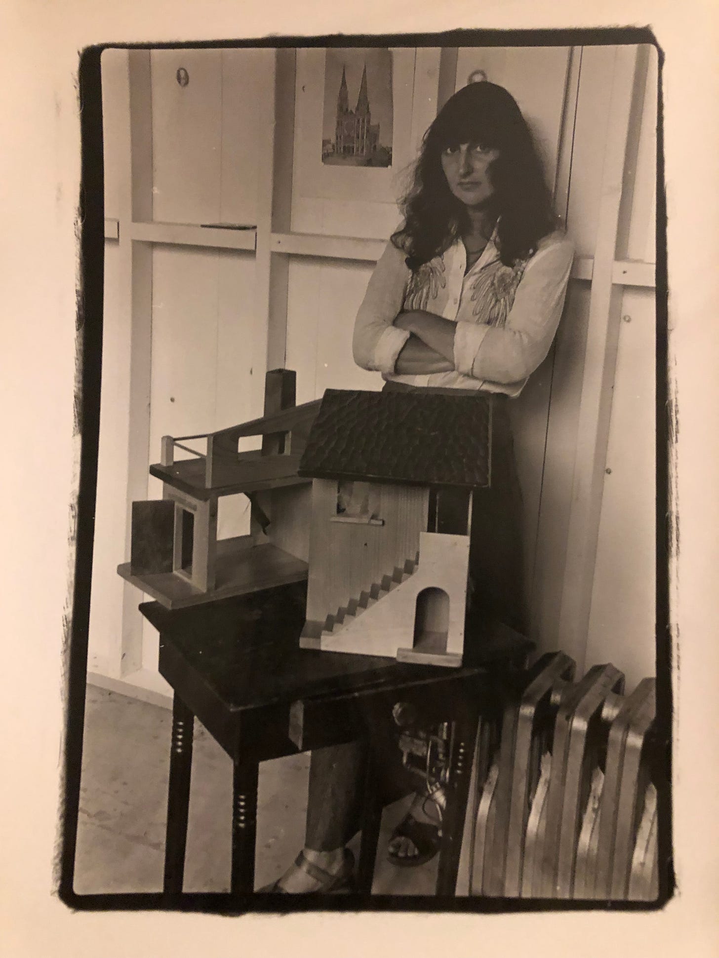 A woman with a petulant look stands with arms crossed leaning against a wall behind a dollhouse on a table.