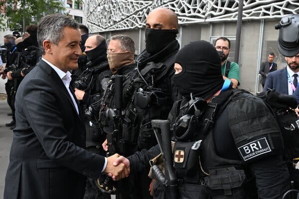 Gérald Darmanin, in a dark suit and tie, shakes hands with a line of police officers in black uniforms and balaclavas covering their faces. 