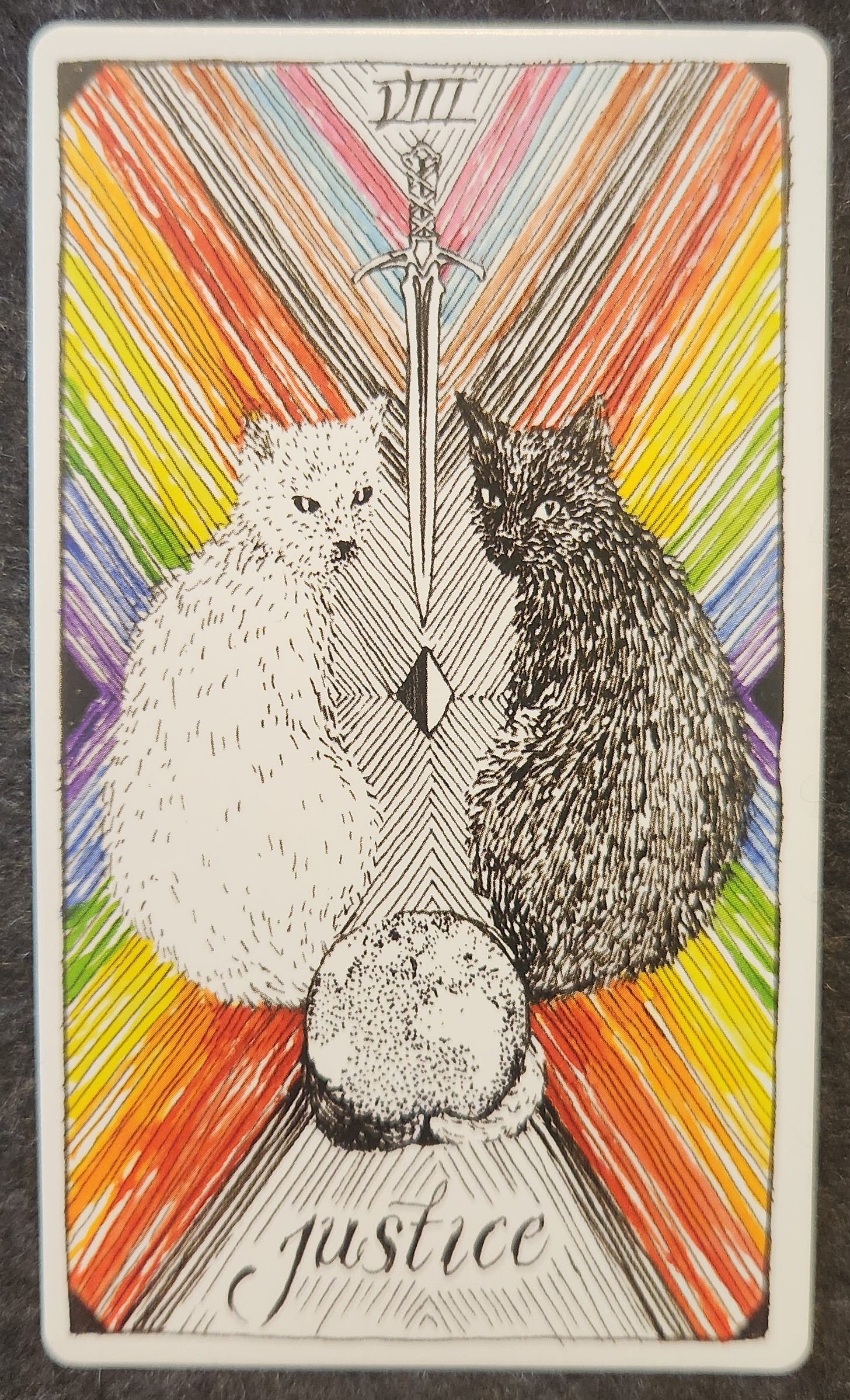 A tarot card labelled XI justice. Two cats, one black and one white, are sitting with their faces turned to the viewer. Between them is a sword with the blade pointed downwards towards a small square shape that is half white and half black. Their tails are wrapped around a round object that looks similar to a black and white globe. 