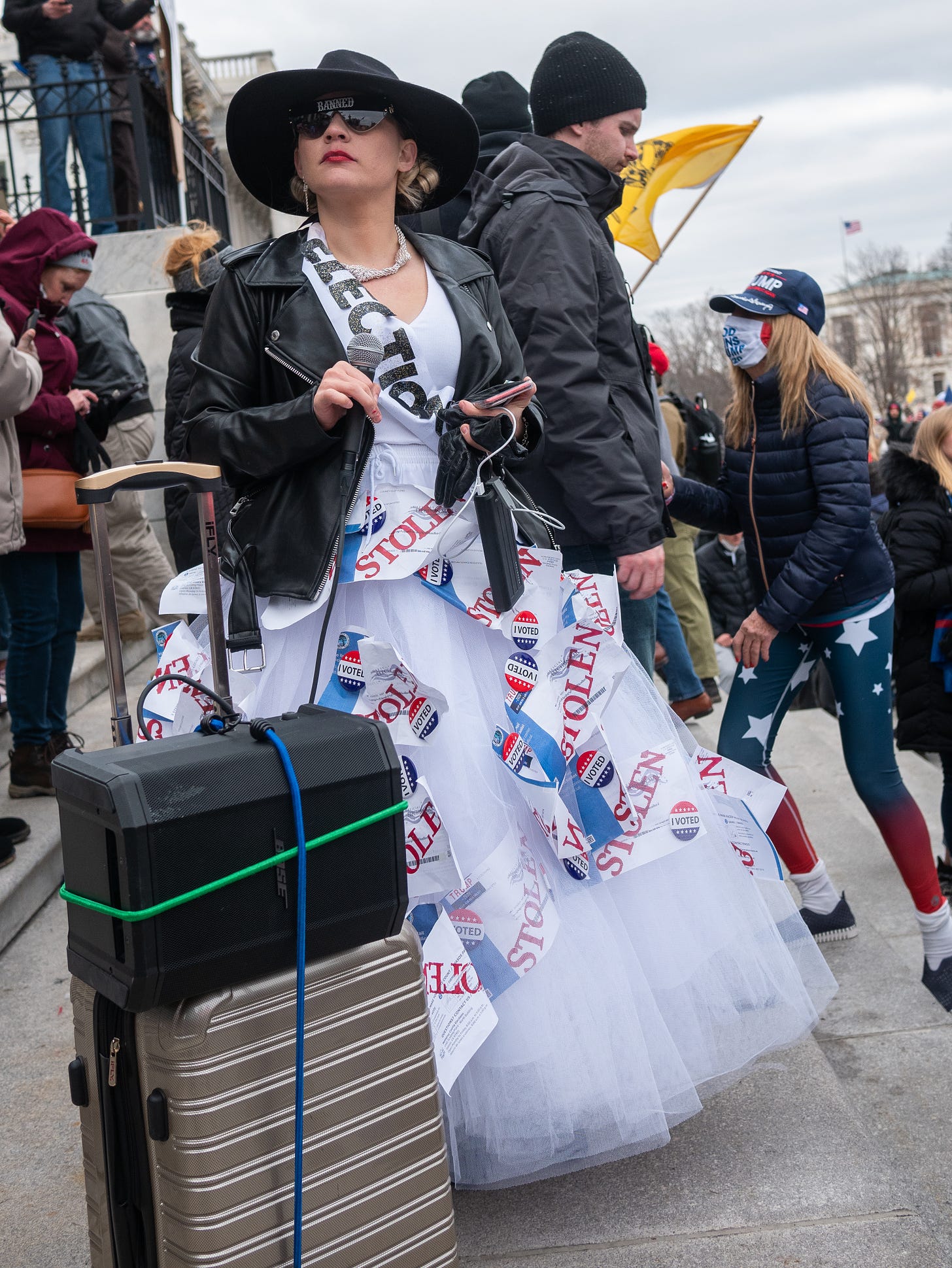 A young woman stands on the steps of the US Capitol on Jan 6, 2021 with an improvised speaker system (a bootbox sized speaker bungied to the top of hard-sided silver carry on luggage). The woman is wearing a long white dress with a big chiffonlike poofy skirt. She is wearing a black leather jacket, wide-brimmed black hat, dark oversized sunglasses, and red lipstick. Blonde curls spill from beneath the hat. She wears a white sash with the word "ELECTION" on it. From the waist down, pieces of paper with the word "STOLEN" in red are affixed to the dress, along with red, white and blue "I VOTED" buttons. She is holding a microphone in her right hand and a phone with a batter bank in her left hand, as if she is preparing to read. She gazes off into the disance as people walk behind her.