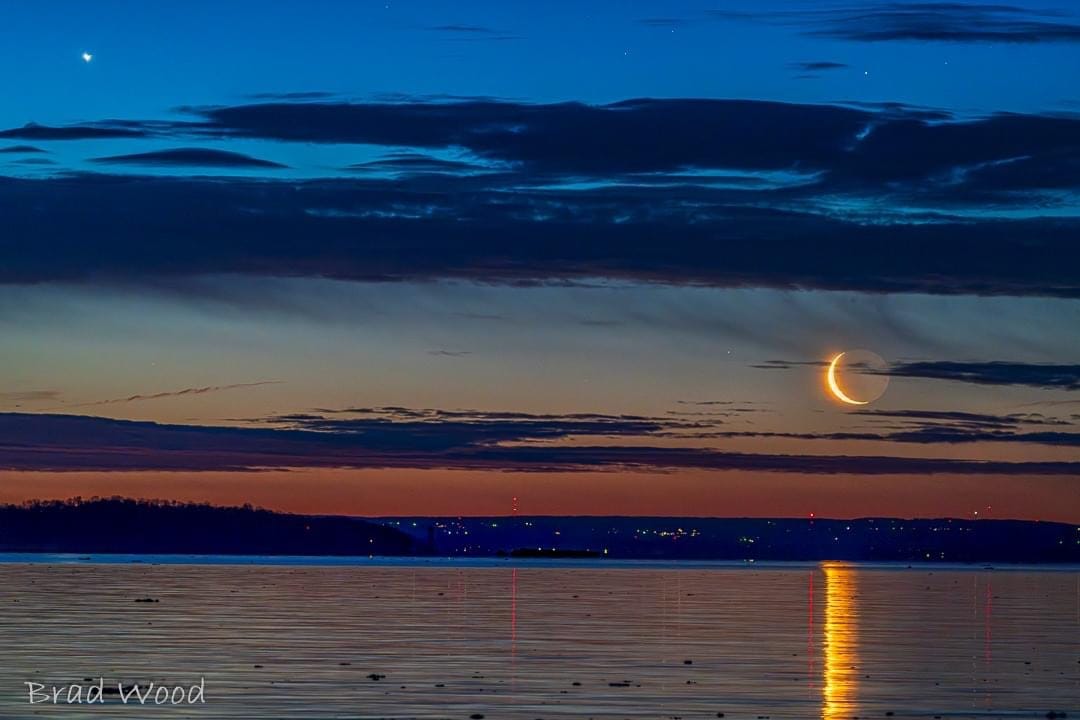 Yellow sliver quarter of a moon shines brightly and reflects on the water below the dark shoreline onto Lake Erie at Port Abino, Ontario in what looks like a combination of a sunset and moonrise.  Although the crescent moon shape looks like it is resting at the left edge on the sun because of the brightness and length of the reflection on the Lake Erie water, it is really the faint shadow of a full moon. The sky is also a dramatic layering of reddish sky, blue-black clouds, light blue sky strip, ending with blue-black clouds with slivers of royal blue to turquoise sky. Photo was taken by Brad Wood and posted on Facebook.